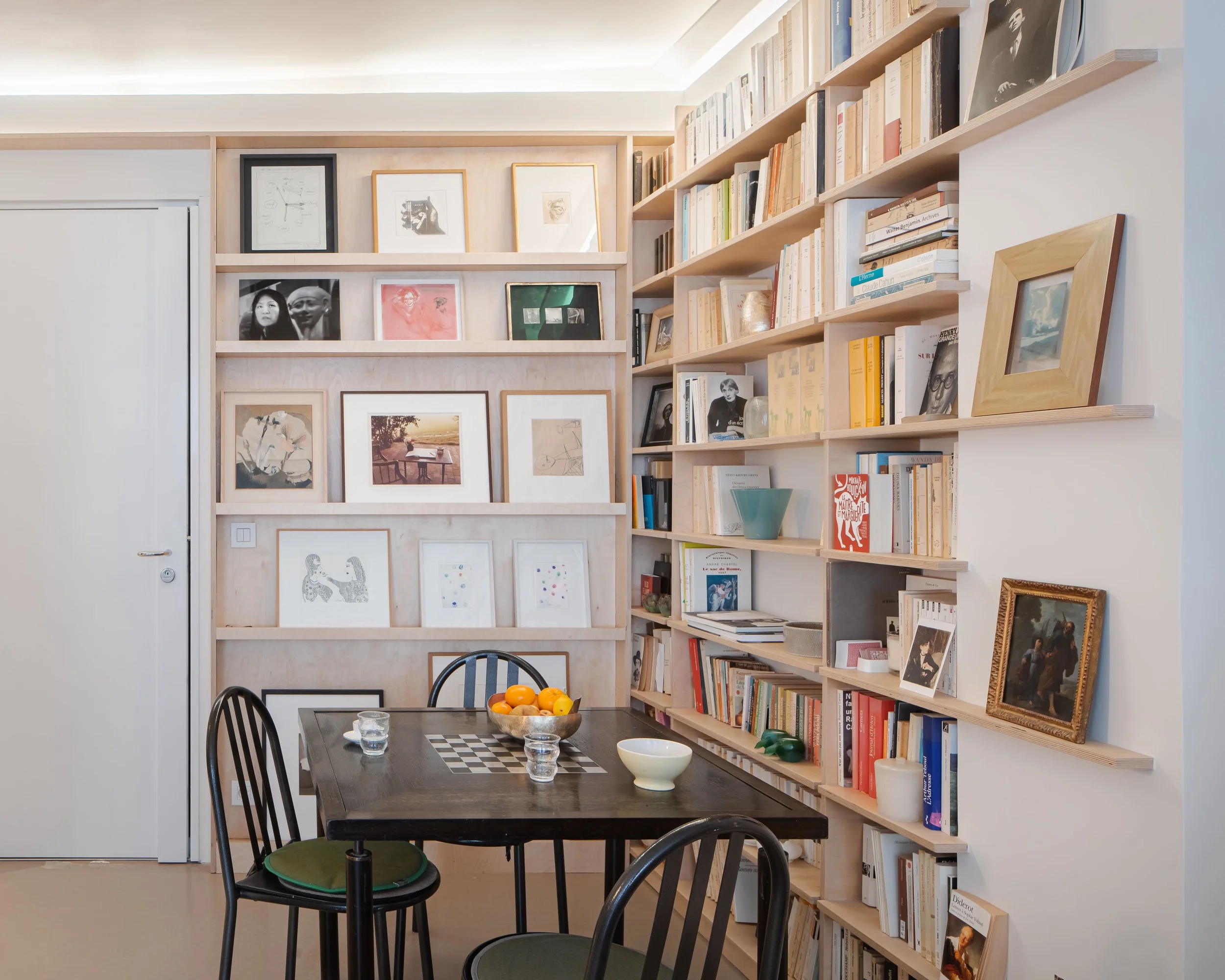 Salle à manger avec une table en bois noir, quatre chaises noires avec coussins verts, et des étagères en bois remplies de livres et de tableaux, devant un mur blanc.