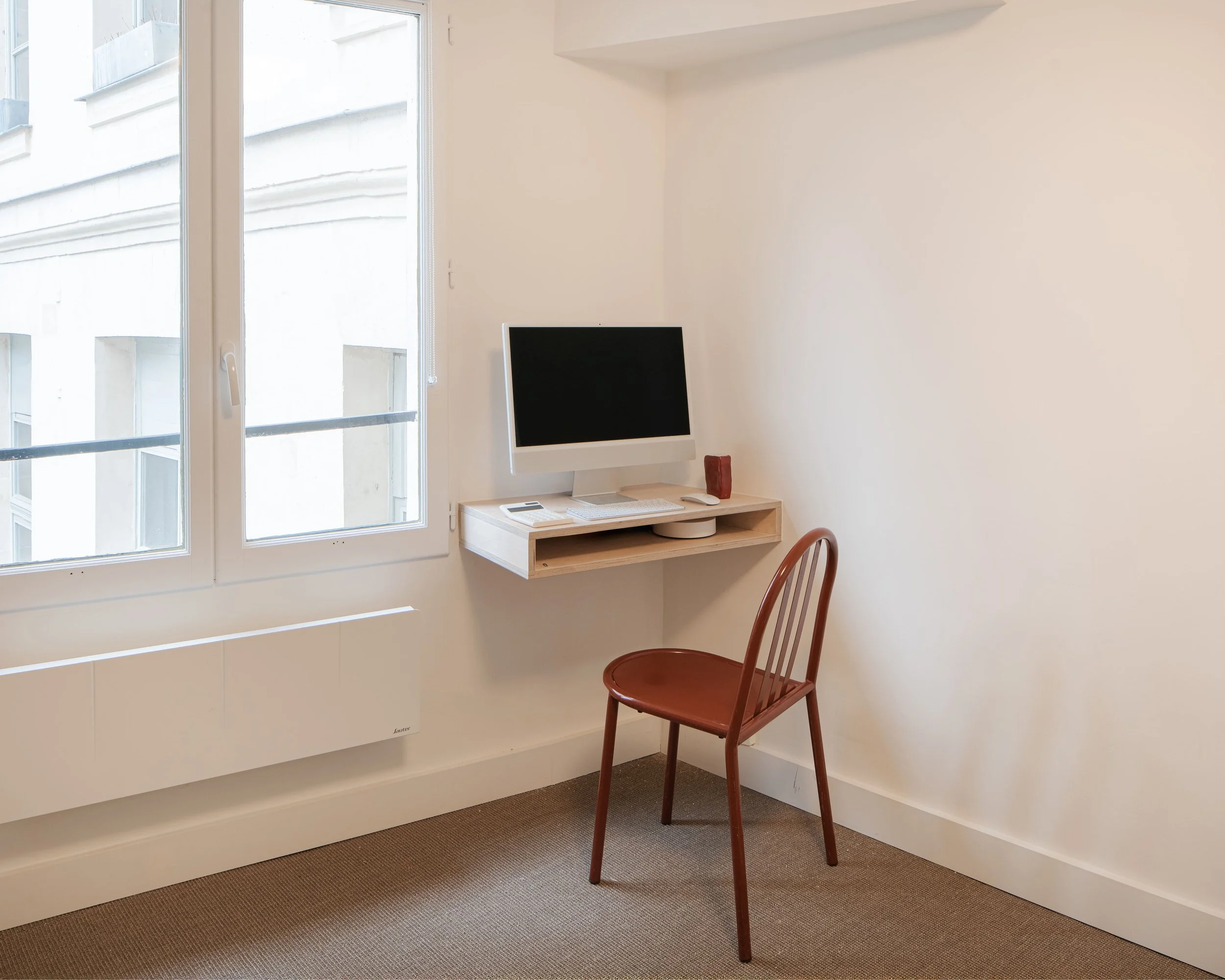 Un bureau minimaliste avec une chaise en bois rouge, un ordinateur blanc sur un bureau mural en bois clair, une fenêtre avec vue sur un bâtiment, et un tapis beige au sol.