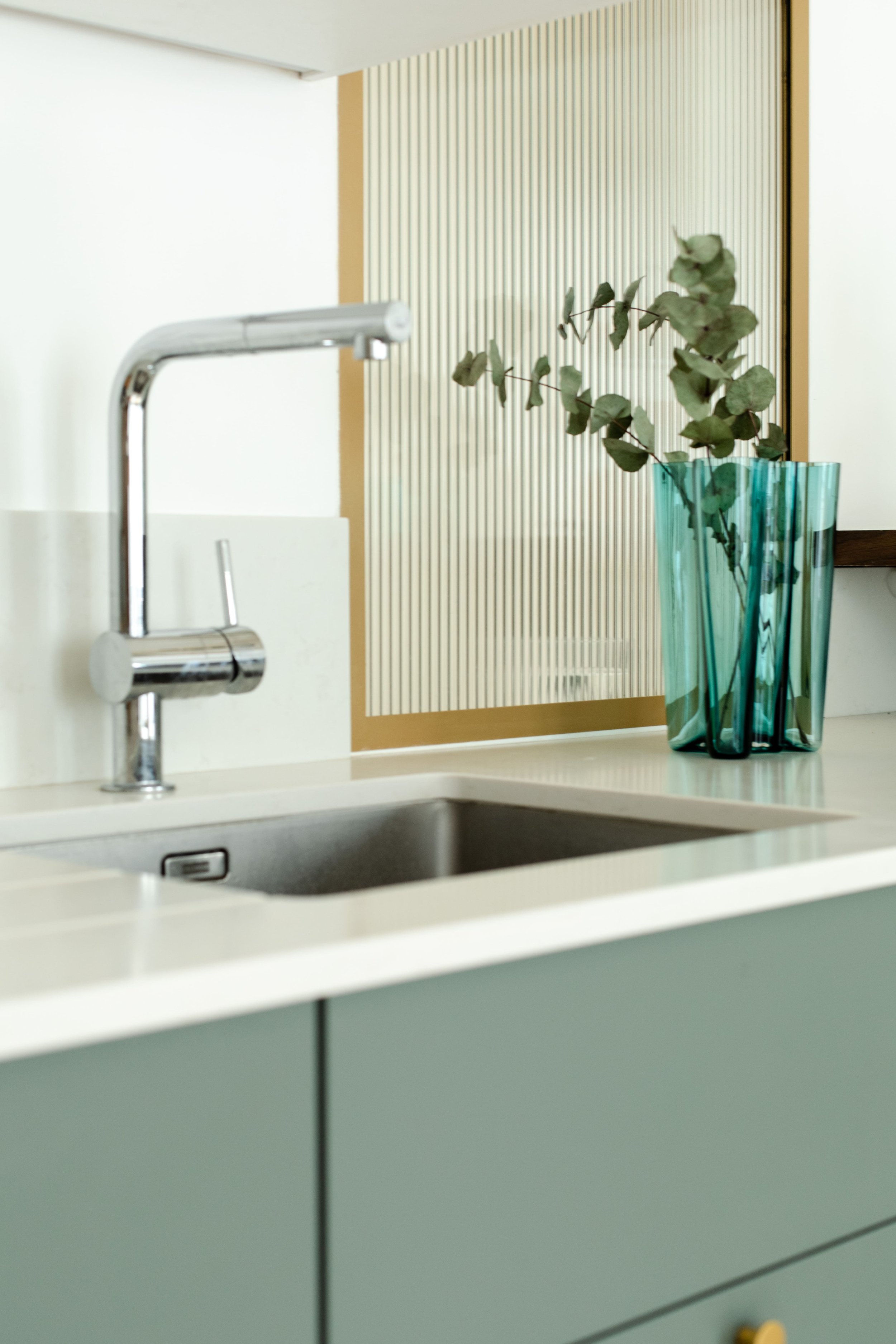 Évier de cuisine avec robinet en chrome, vasque en acier inoxydable, comptoir blanc, porte de placard vert sauge, vase en verre turquoise avec des branches de feuillage vert.
