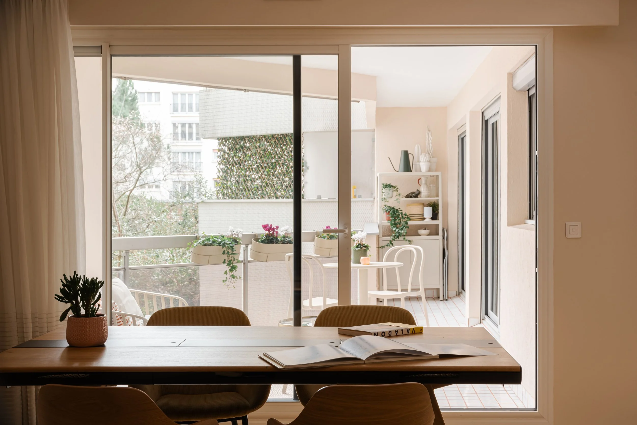 Intérieur d'une salle avec une table en bois, un livre ouvert, un livre ferme, et une plante en pot, regardant sur un balcon avec des chaises, une table, des plantes et des étagères, dans un appartement moderne.
