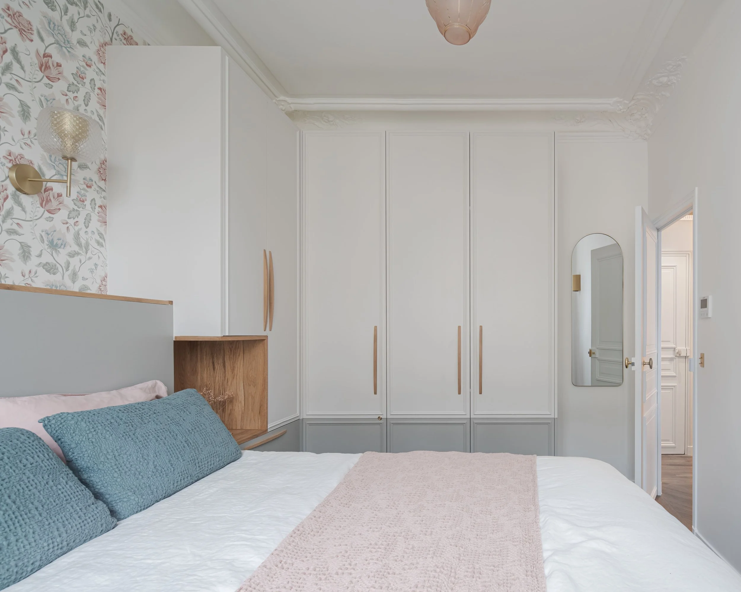 Chambre à coucher moderne avec lit, coussins bleus, armoires blanches, miroir mural et porte ouverte vers un autre pièce.