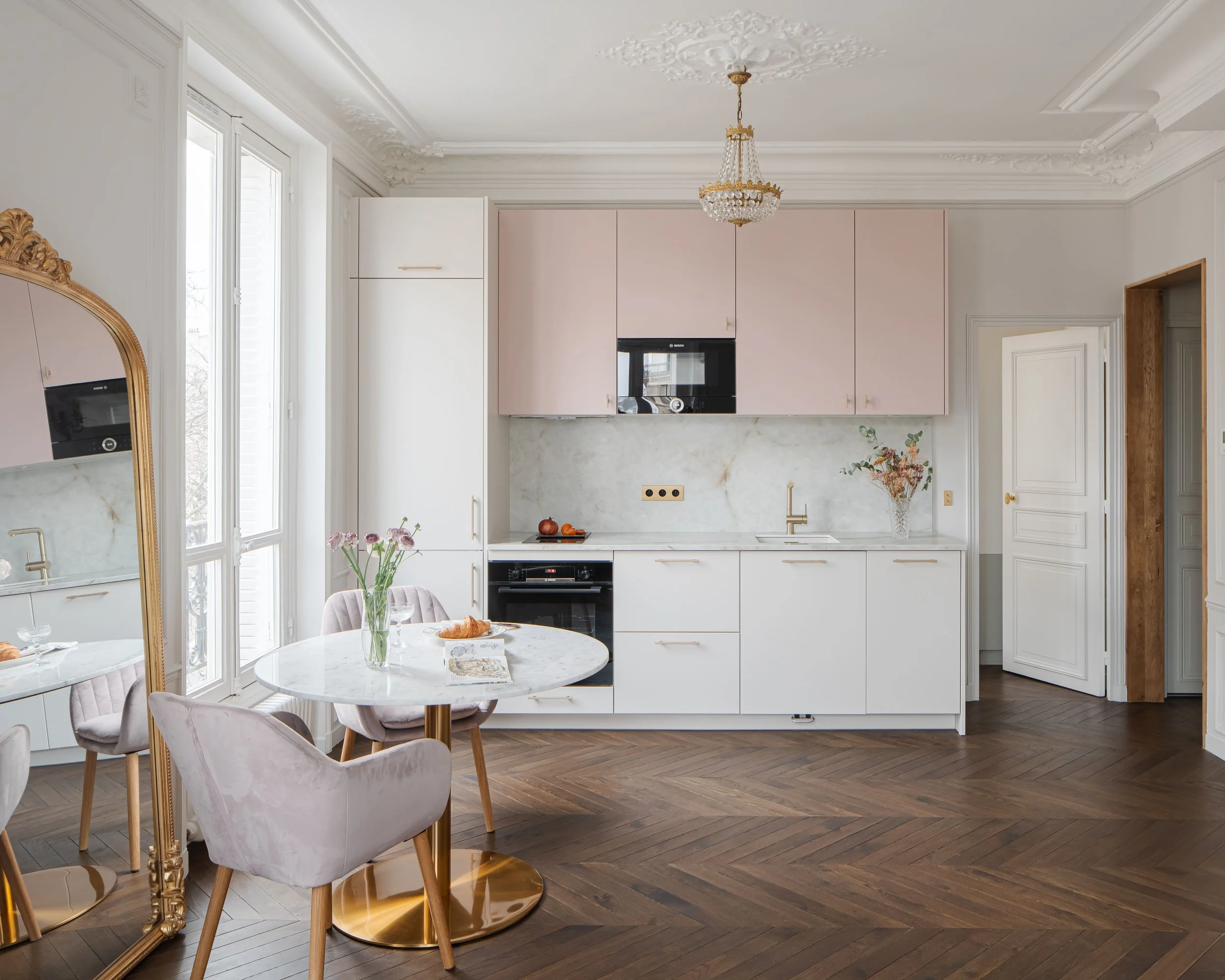 Cuisine moderne avec des placards rose pastel, une table ronde en marbre avec des chaises en velours rose clair, un chandelier et un grand miroir doré.