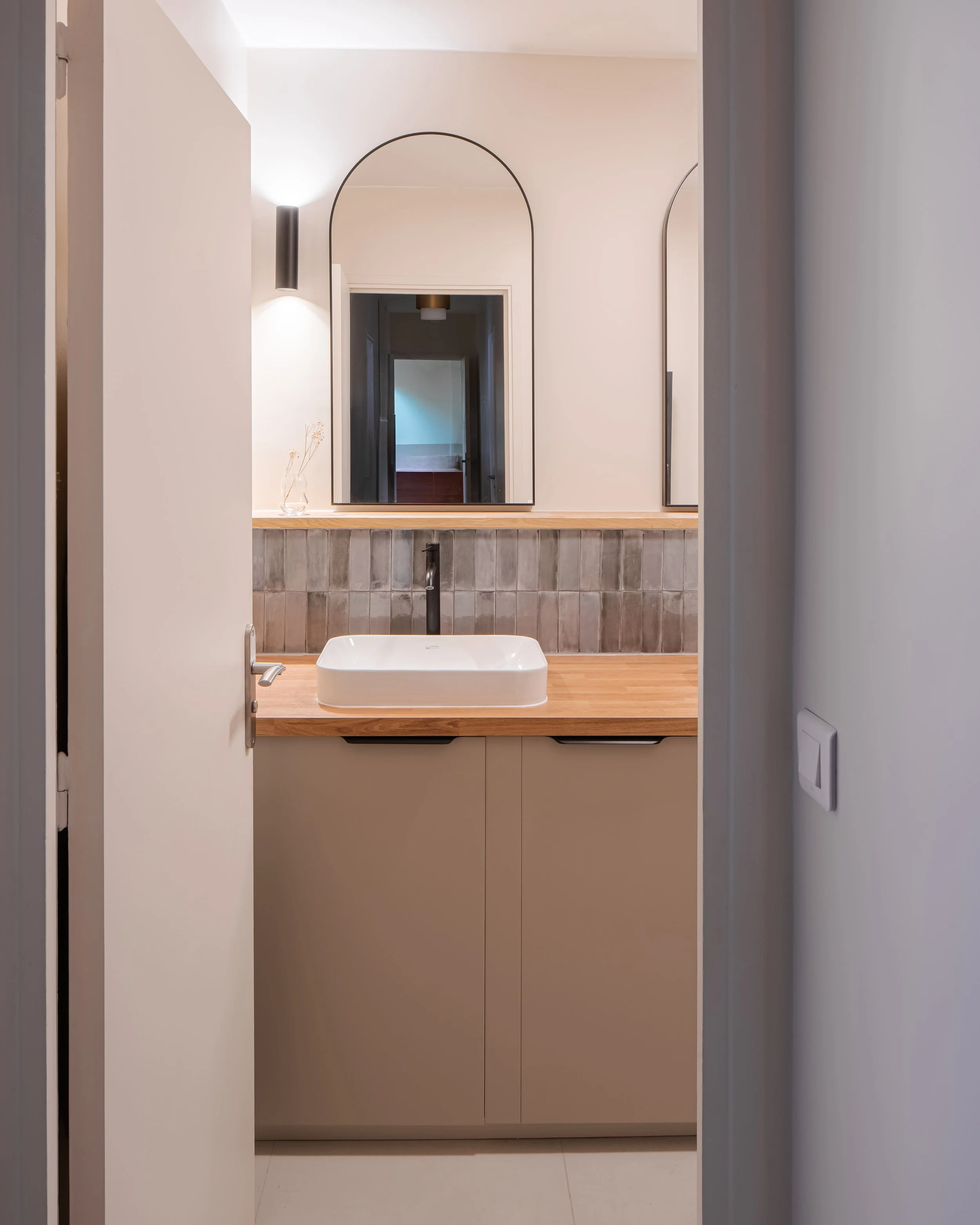 Intérieur d'une salle de bain avec un lavabo carré en céramique sur un plan de travail en bois, deux miroirs ovales et une lumière murale.
