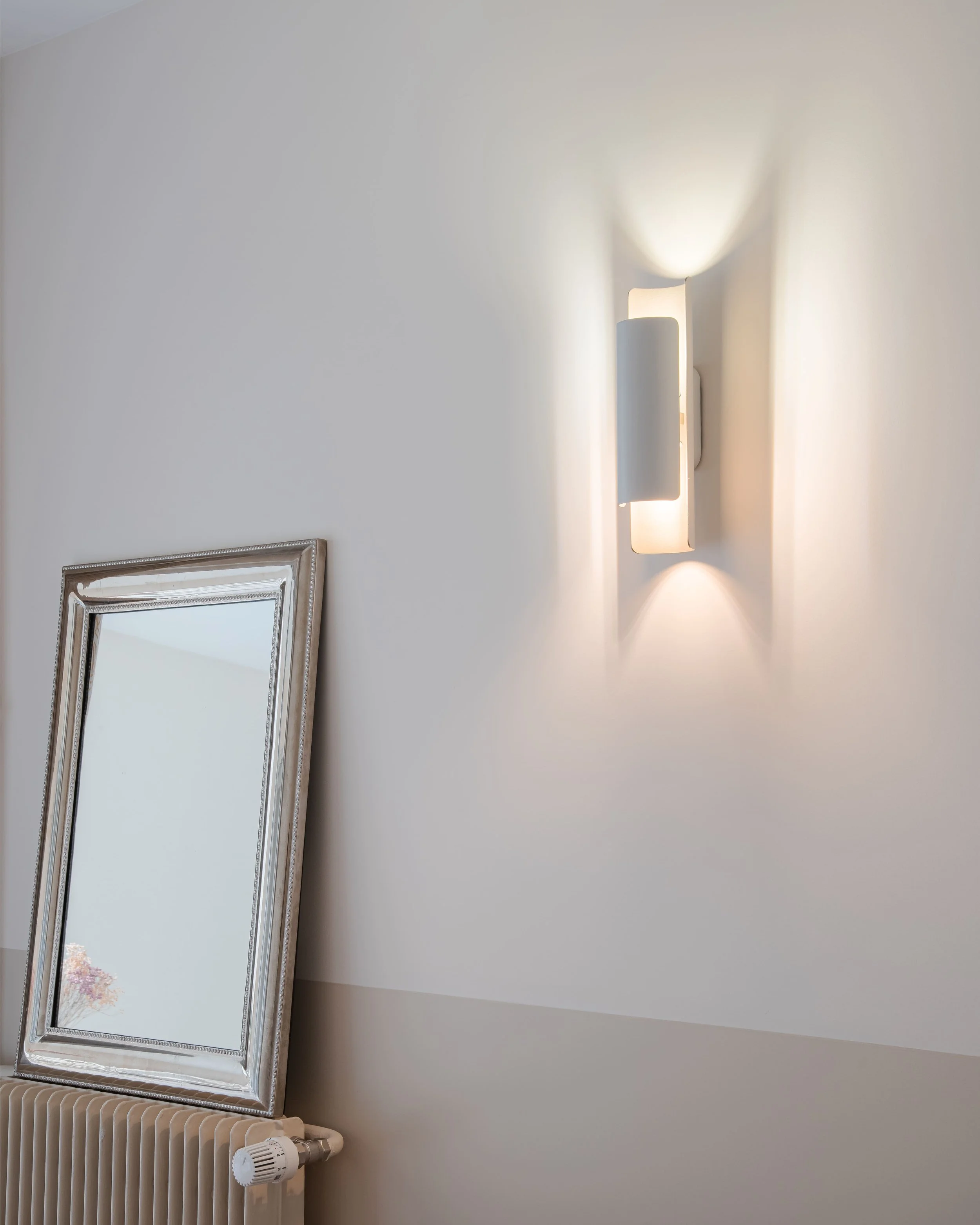 Un miroir avec un cadre argenté reposant sur un radiateur blanc, à côté d'une applique murale blanche allumée, sur un mur blanc.