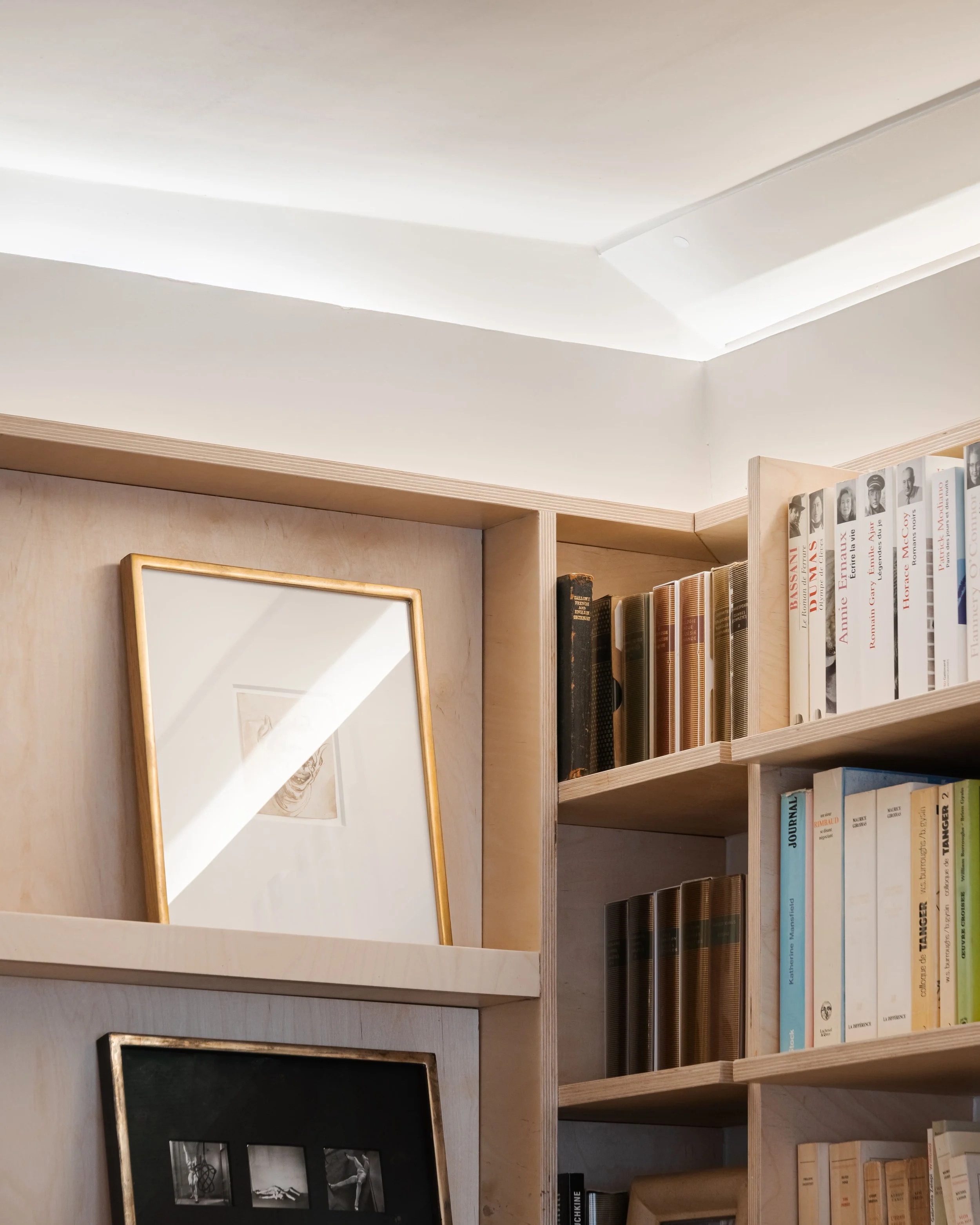 Une bibliothèque en bois clair avec des livres et des cadres photo, sous un plafond blanc.