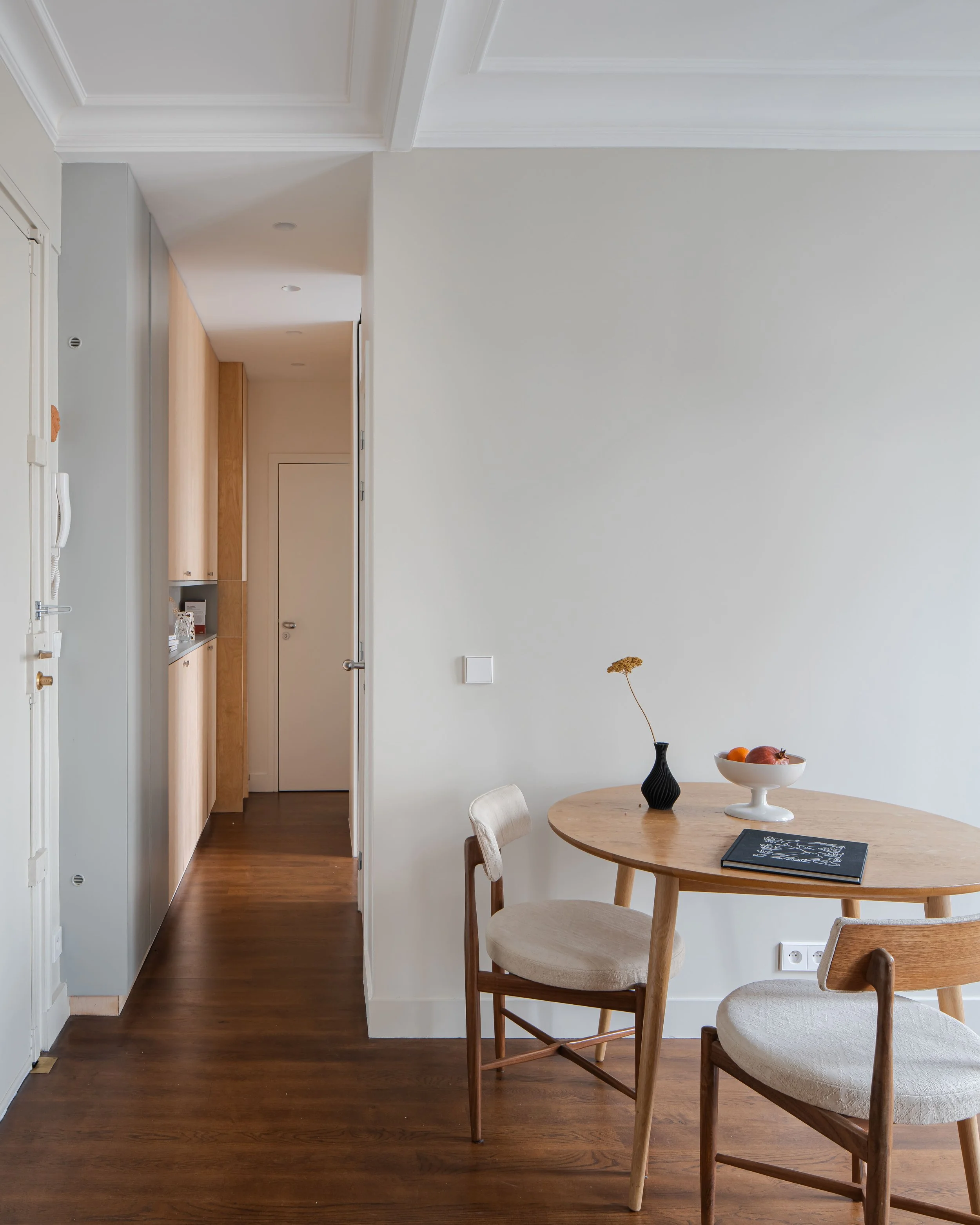Intérieur d’un appartement moderne avec une table ronde en bois, deux chaises en bois avec coussins, une carafe noire avec une fleur, un bol blanc avec des fruits, sols en bois foncé, murs blancs, et porte menant à une cuisine