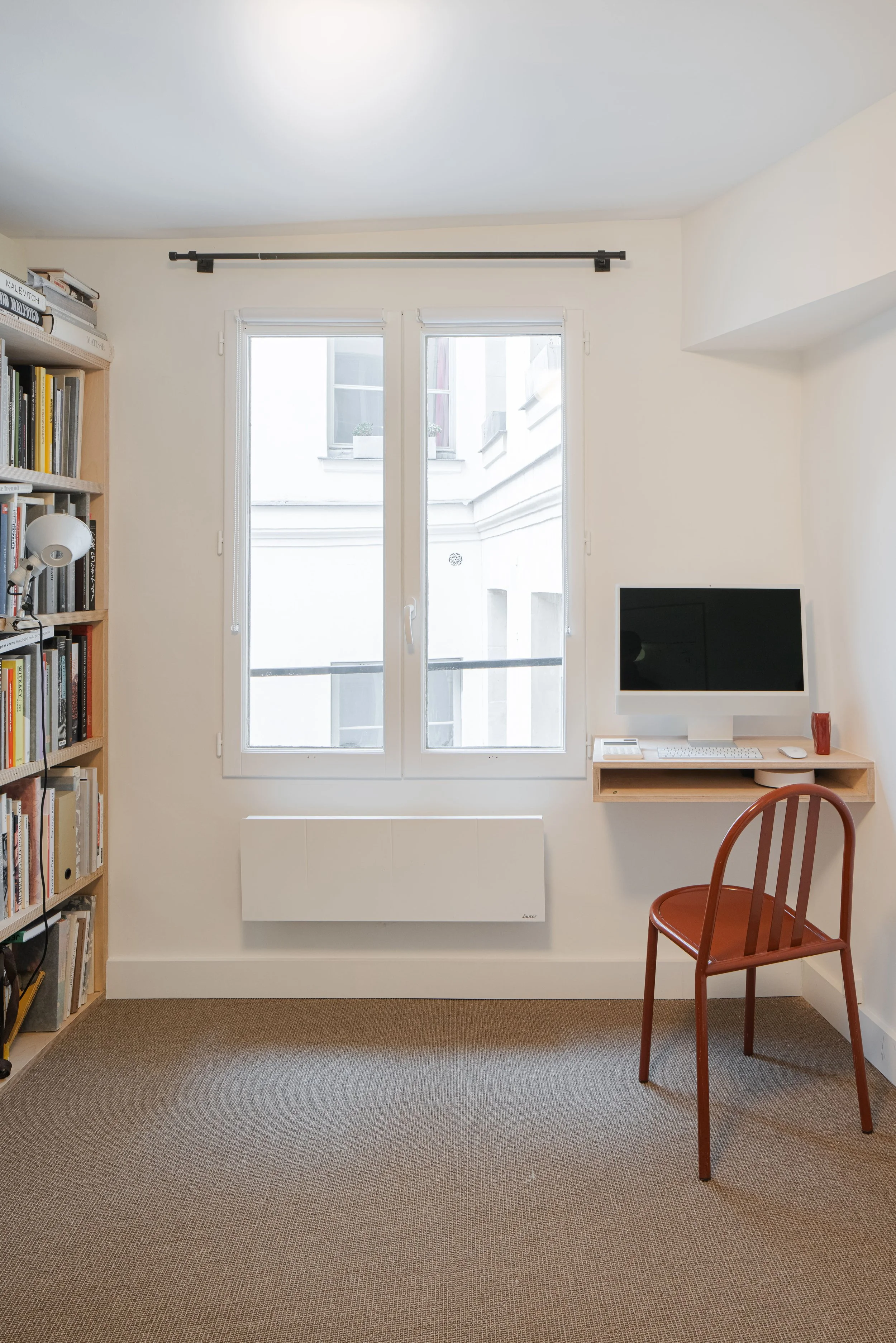Petite pièce avec une bibliothèque en bois à gauche, une fenêtre centrale, un radiateur blanc dessous, un bureau flottant blanc à droite avec un ordinateur, clavier, souris et une tasse ou vase rouge, et une chaise en bois rouge devant le bureau.