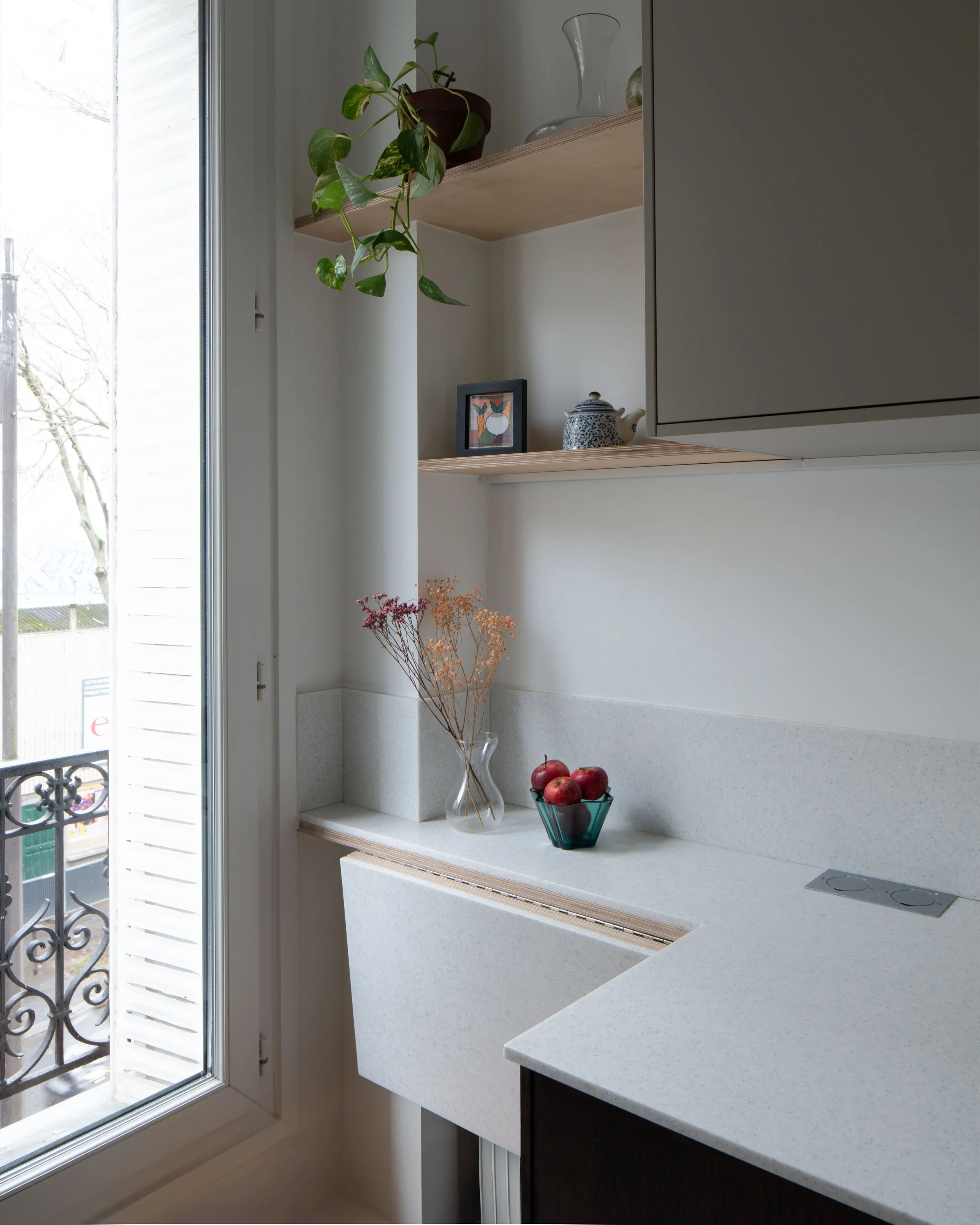 Cuisine moderne avec fenêtre donnant sur un balcon, étagères en bois avec plantes, vase avec des fleurs séchées, bol de pommes, et objets décoratifs