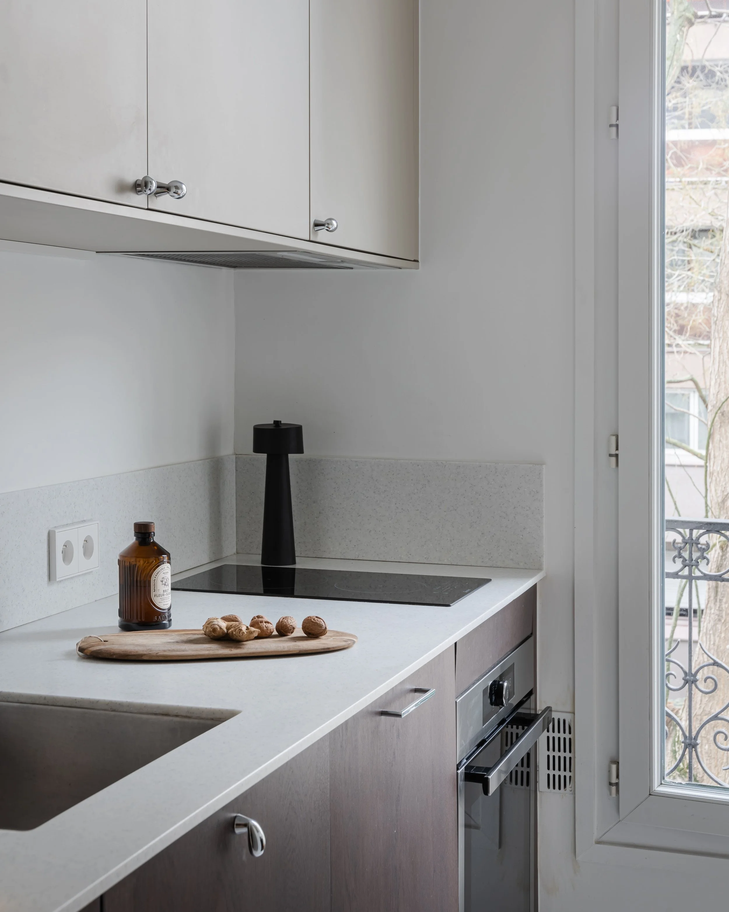 Cuisine moderne avec plan de travail en pierre, bouteille en verre, noix, plaque de cuisson électrique, placard en bois clair, grande fenêtre avec vue sur des arbres.