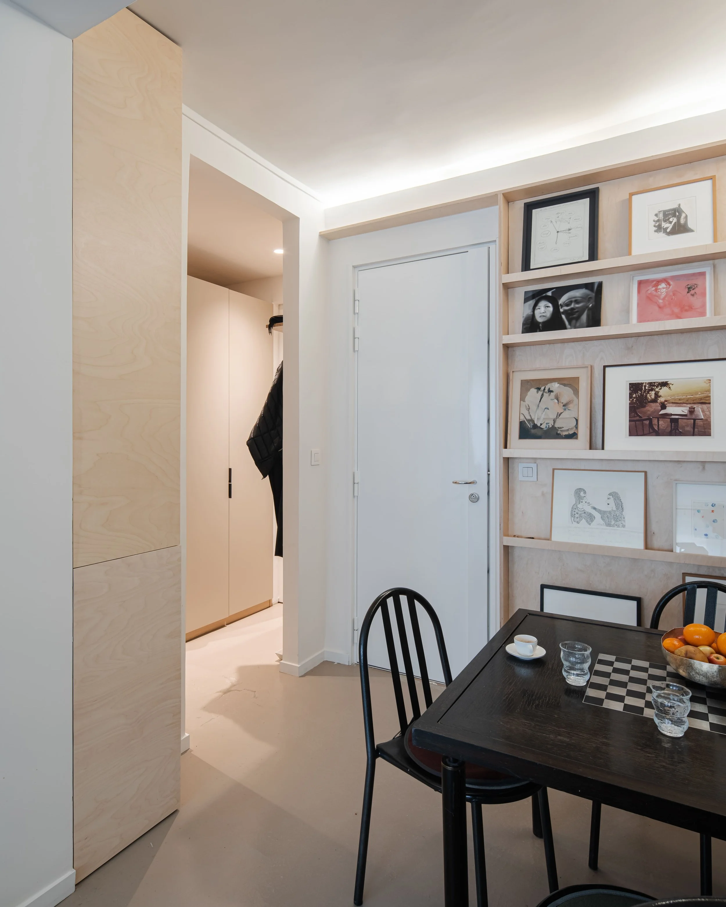 Salle à manger avec un mur de photos encadrées, une table noire avec des chaises, un bol de fruits et des verres, et une porte blanche vers une entrée ou un couloir.
