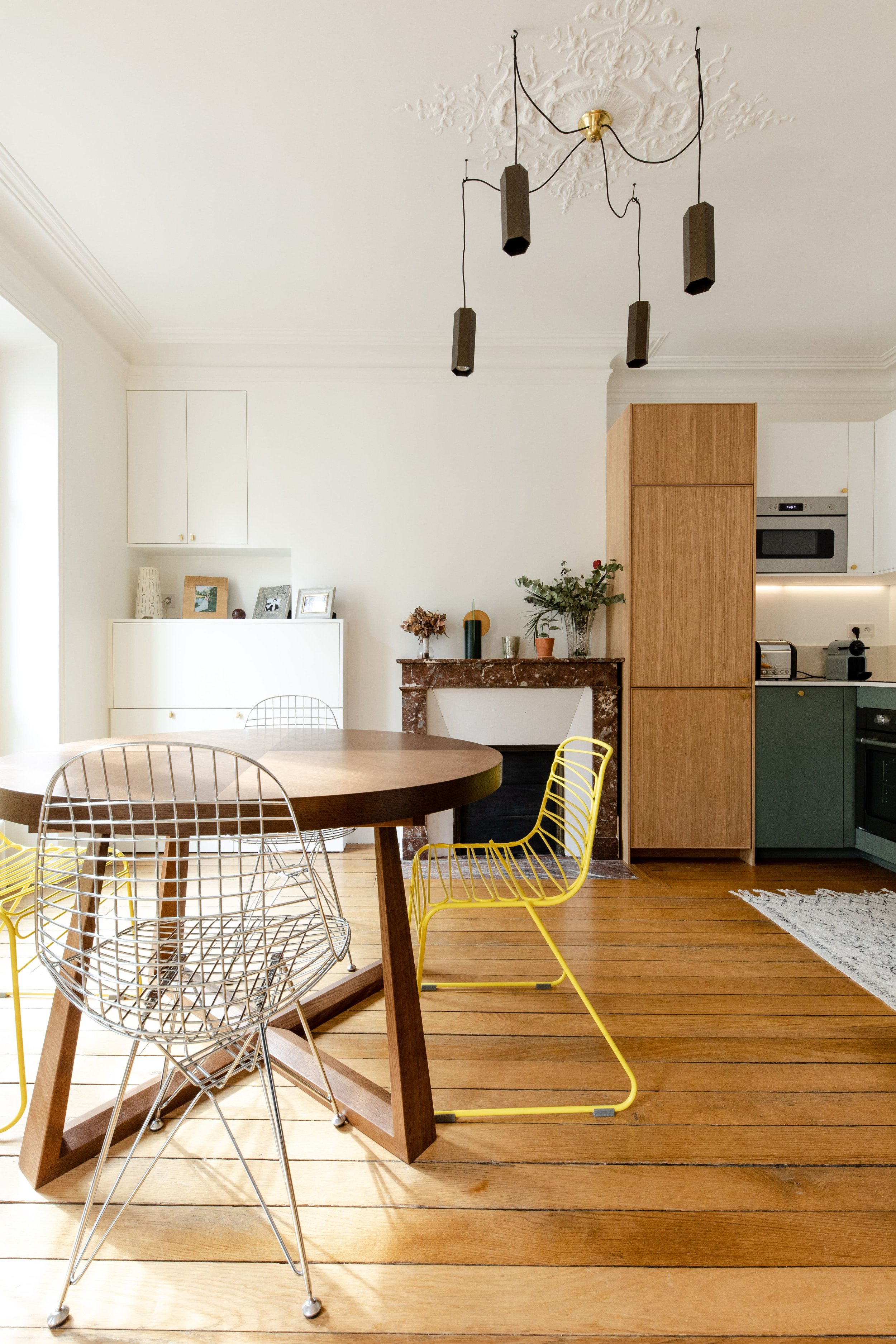 Une cuisine et salle à manger modernes avec un plafond blanc orné de moulures, un lustre noir suspendu, une table en bois ronde avec des chaises en fil métallique, un mur blanc, un meuble blanc avec des cadres photo, une cheminée en marbre avec des plantes, et des armoires en bois clair. Sol en bois.»