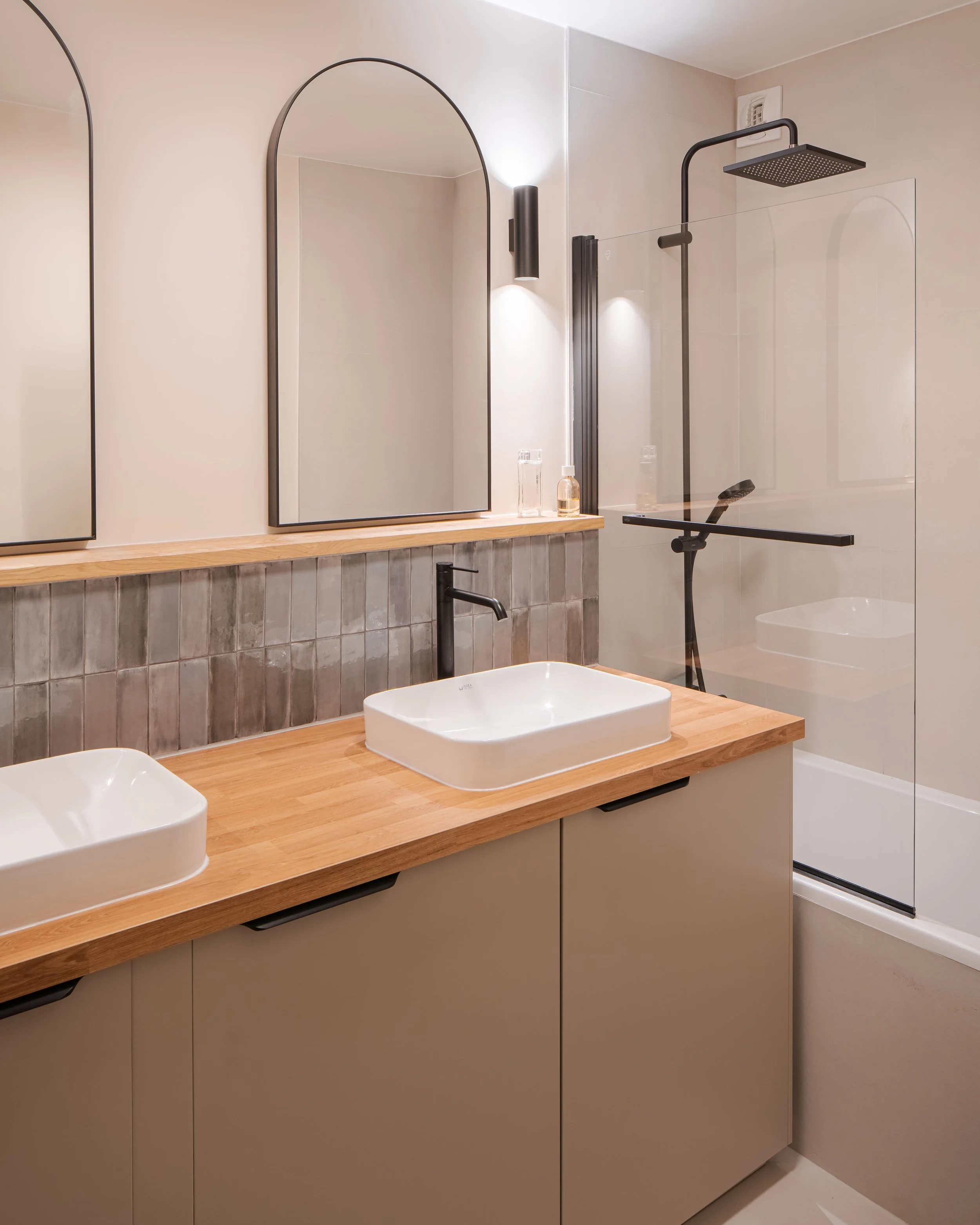Salle de bain moderne avec deux lavabos carrés sur un plan en bois, grands miroirs, douche avec paroi en verre, robinet noir, et décoration minimaliste