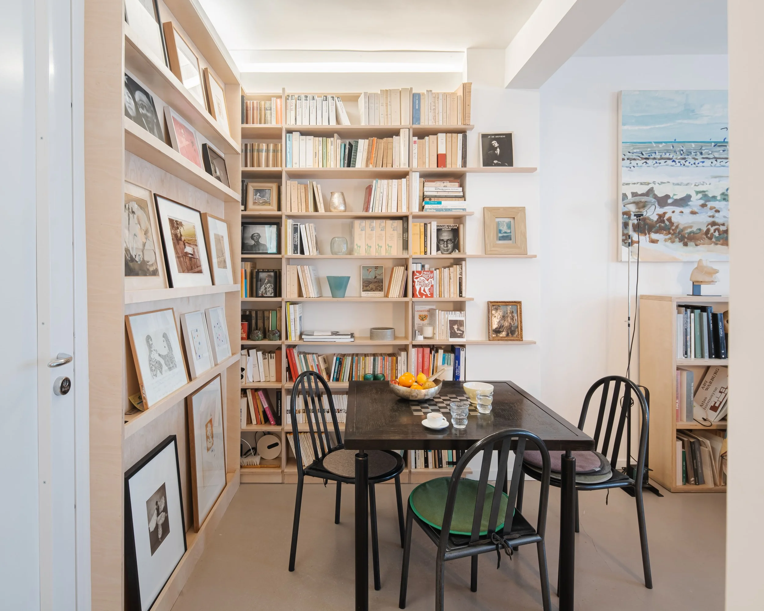 Salle à manger avec un tableau en bois noir, un bol de fruits, 4 verres, assiettes et une tasse sur une table en bois, entourée de 4 chaises. Mur avec étagères remplies de livres et d'objets décoratifs, une œuvre d'art représentant la mer au mur à dr