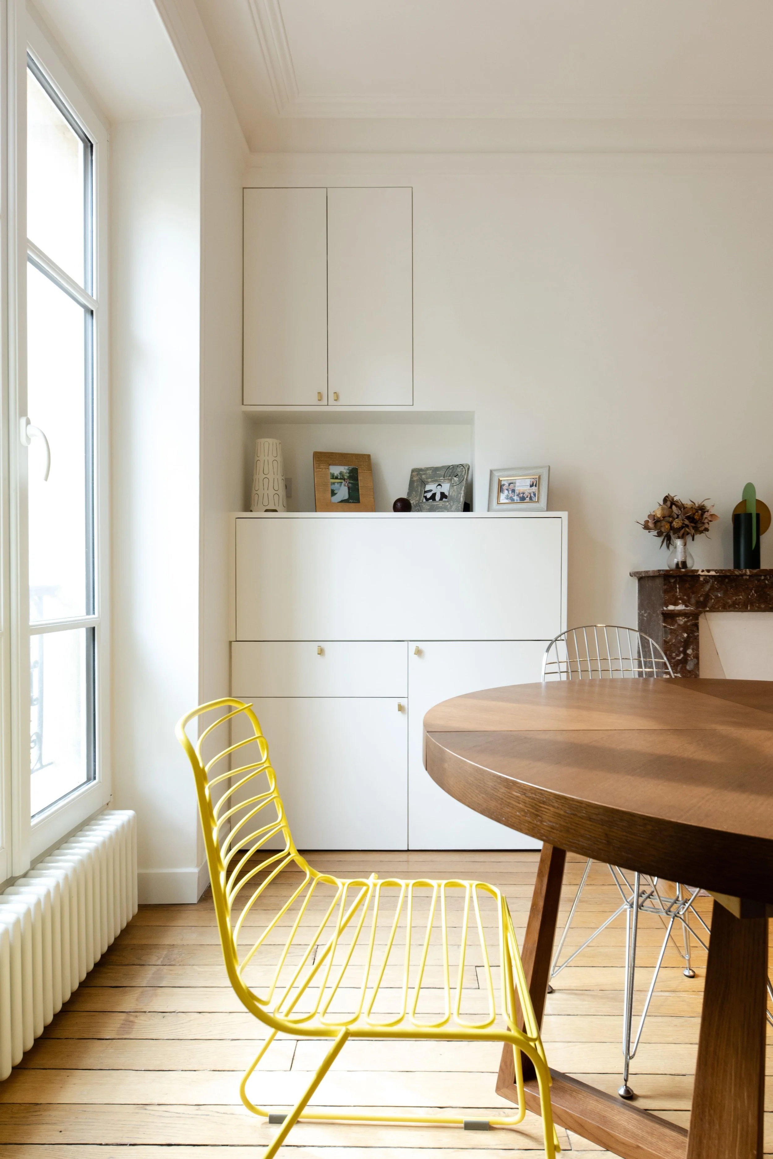 Intérieur d'une salle à manger avec une table en bois, une chaise jaune en métal, une cheminée et un meuble blanc avec des cadres photos et un vase.