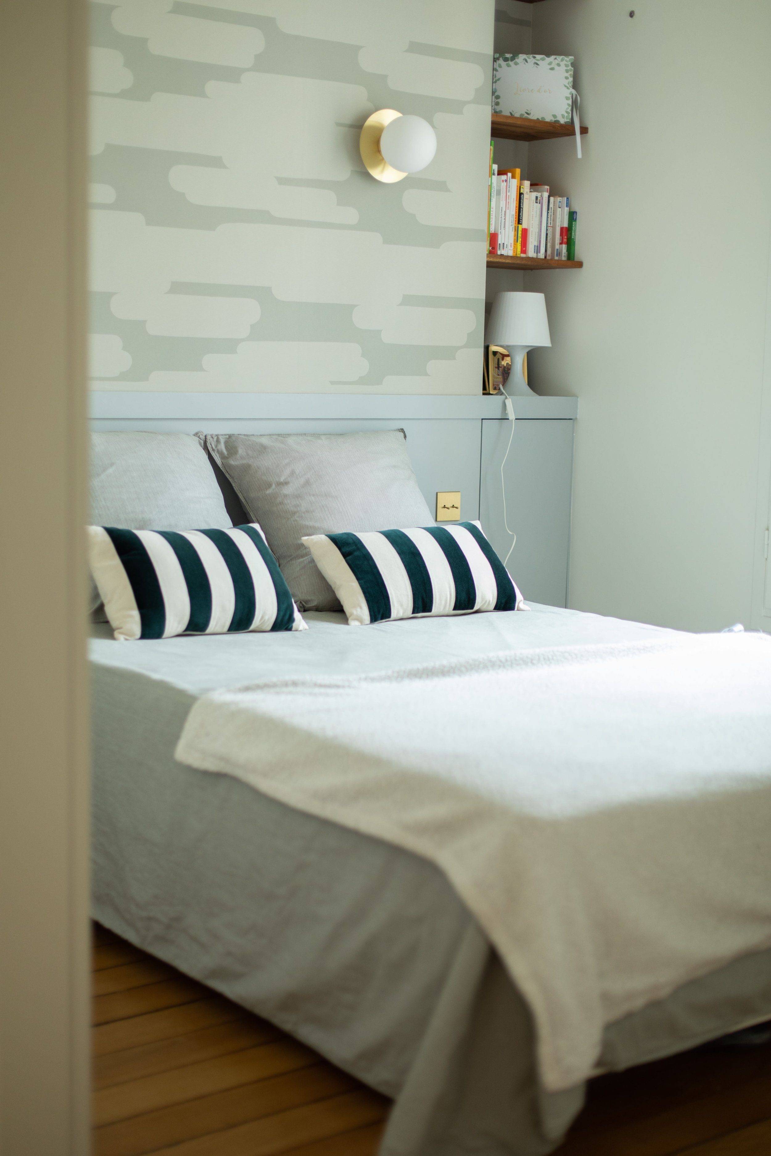 Chambre à coucher avec un lit fait avec une housse blanche, deux oreillers gris et deux coussins rayés noir et blanc. Mur avec une lampe.?a out, étagères en bois avec des livres et une lampe de chevet blanche.