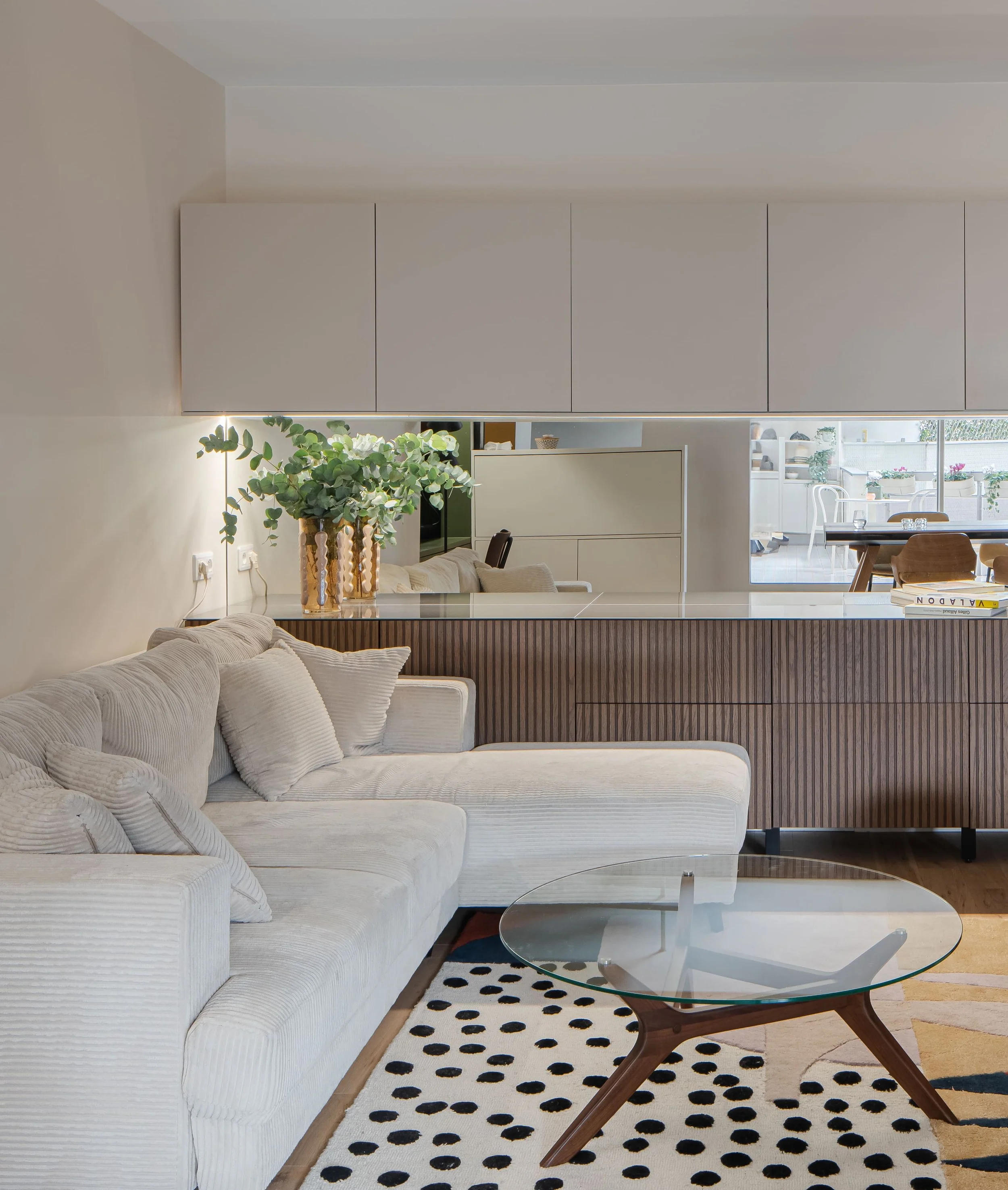 Salon moderne avec canapé beige, tapis noir et blanc, table en verre et cuisine ouverte avec planche en bois et végétaux