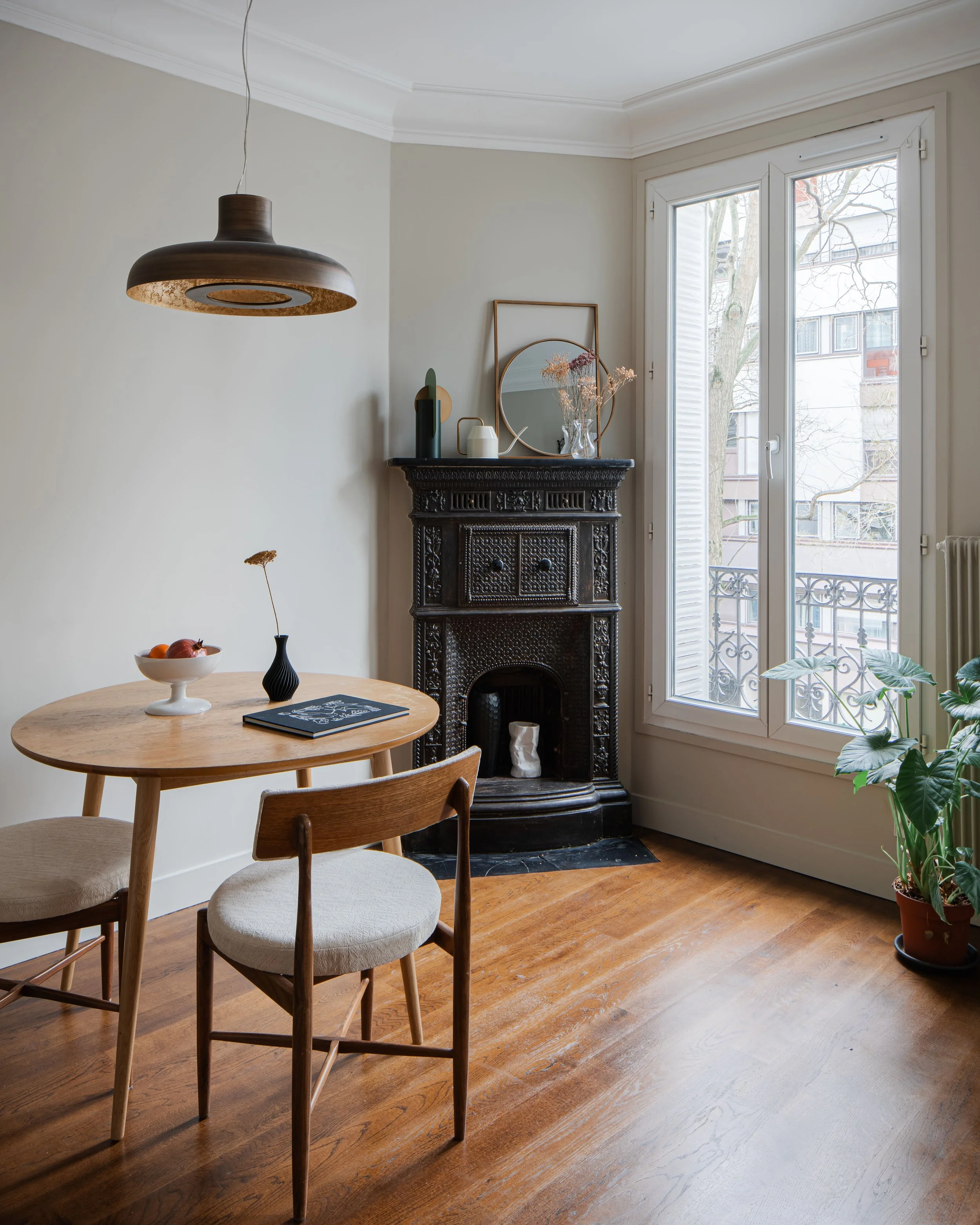 Intérieur d'un salon lumineux avec un balcon, cheminée noire en bois sculpté, table en bois avec deux chaises, pot de fleurs, et décorations modernes sur la cheminée, le tout avec un parquet en bois.