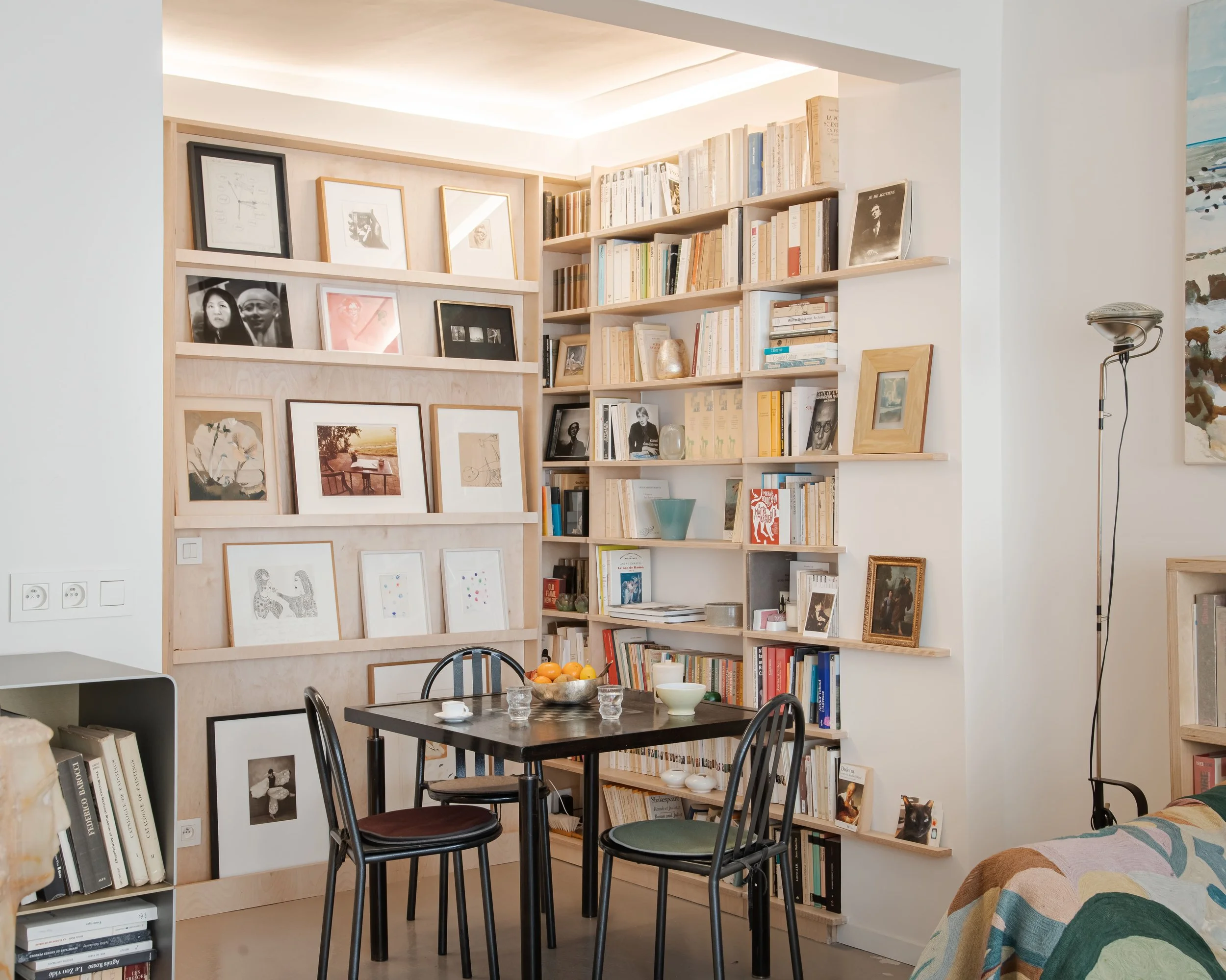 Salle à manger avec table noire, trois chaises, étagères remplies de livres et photos encadrées, canapé avec couverture colorée, lampadaire à gauche.