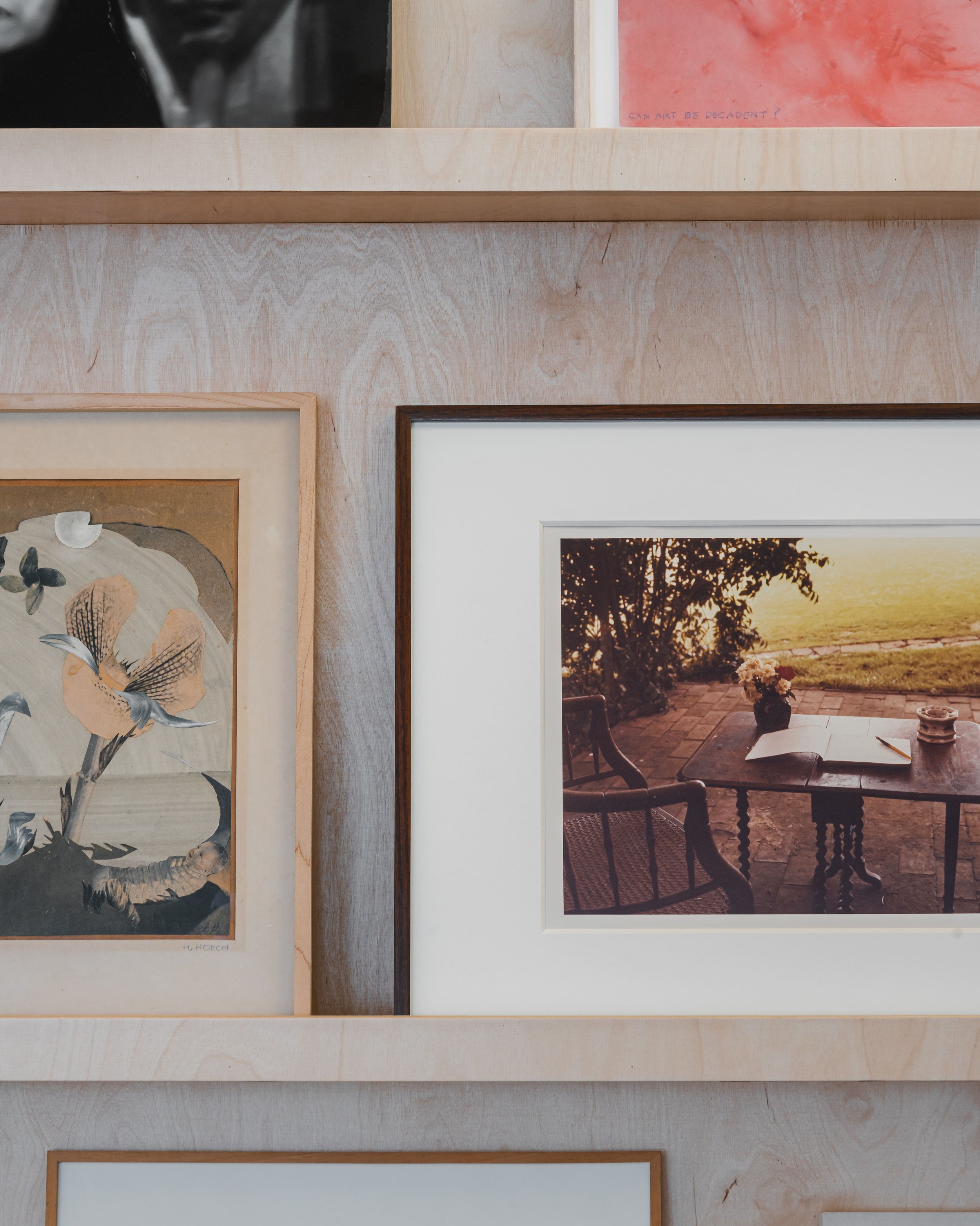 Photographie de deux œuvres d'art encadrées sur un mur en bois clair. La gauche représente un motif floral stylisé et la droite montre une scène extérieure avec une table, deux chaises et la nature au coucher du soleil.