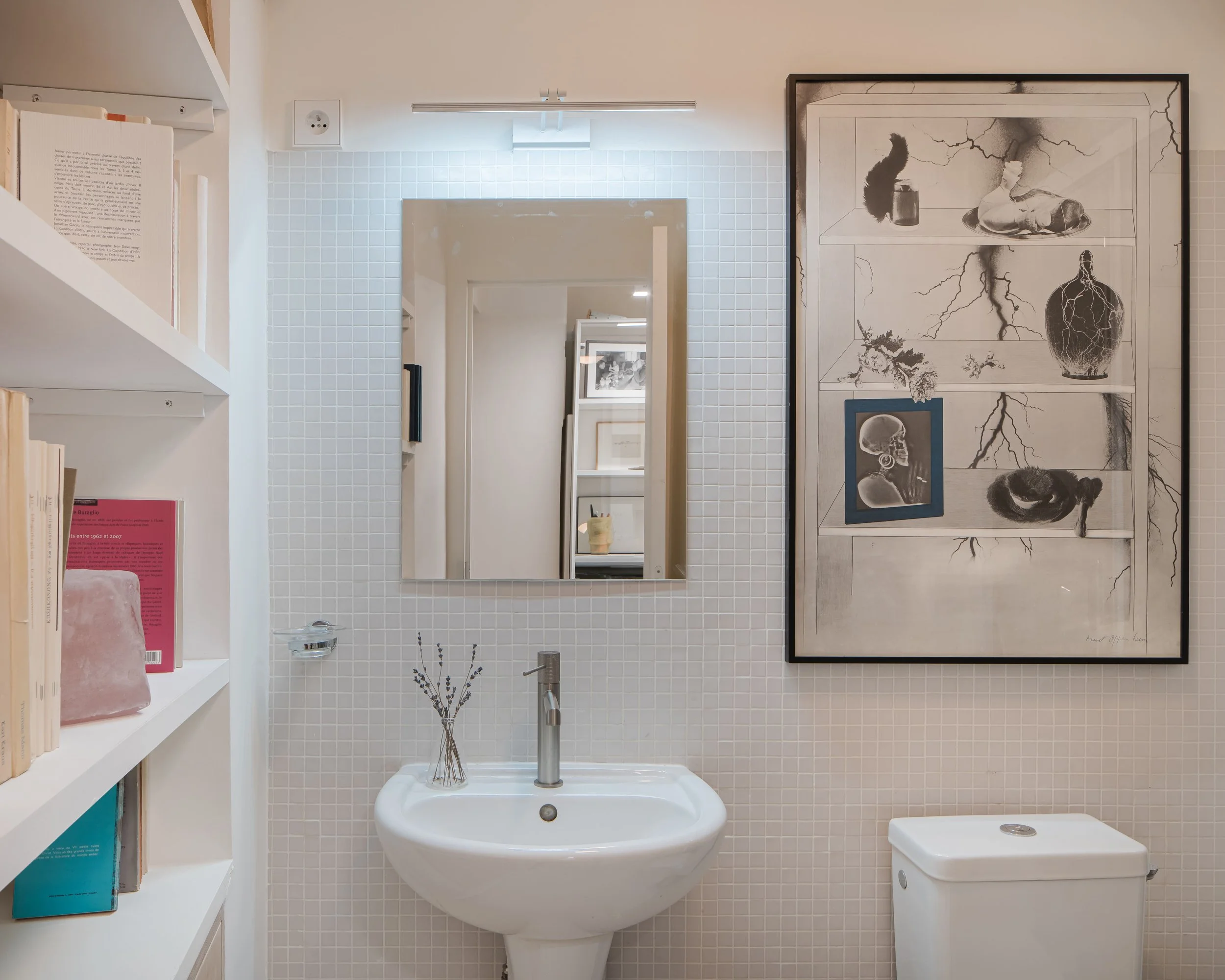 Salle de bain avec un miroir, un lavabo, une étagère avec des livres, un tableau d'art abstrait, et un lavabo blanc avec une petite décoration florale.