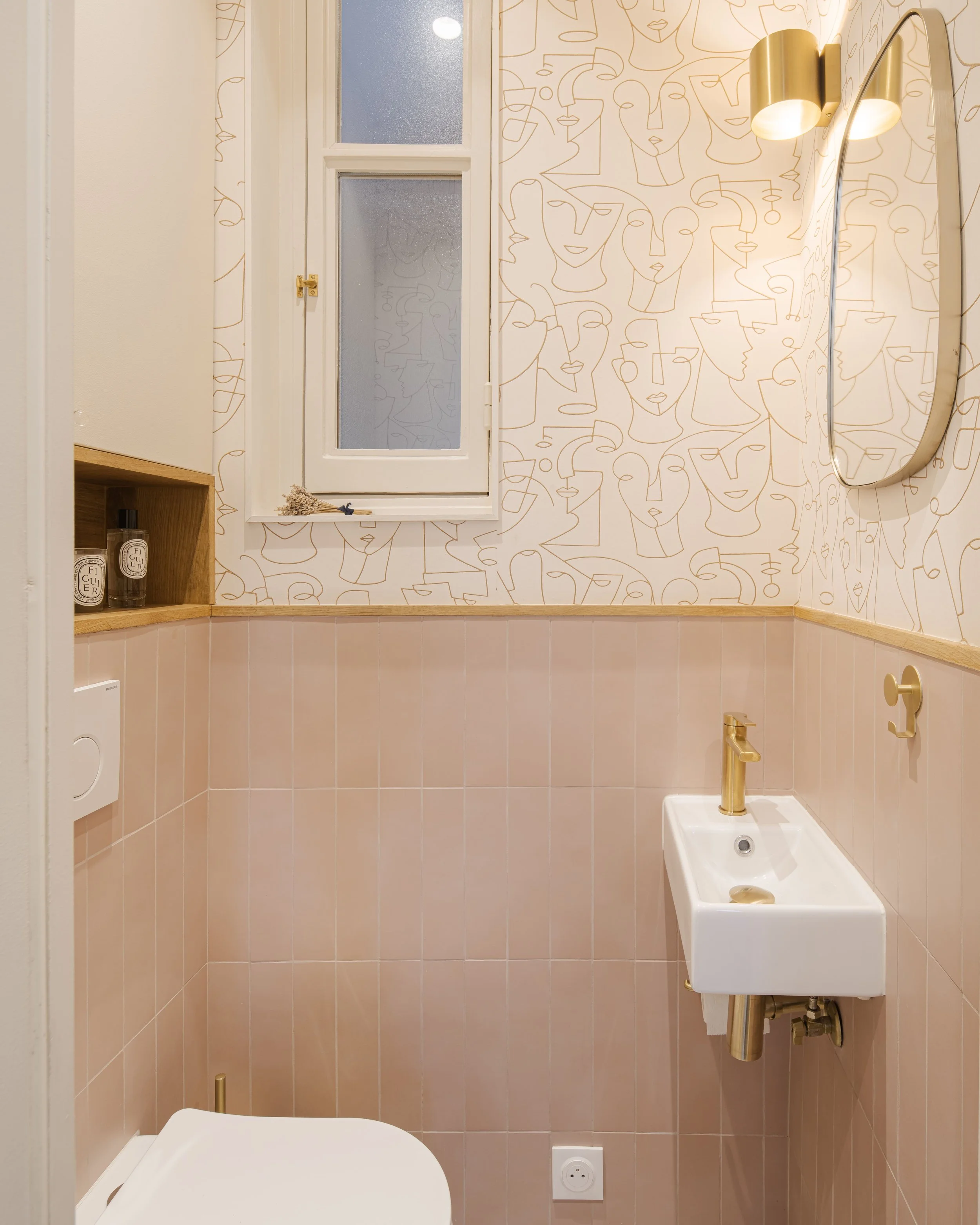 Une petite salle de bain moderne avec papier peint à motifs de sujets abstraits, une fenêtre, un miroir, un lavabo blanc avec robinet en or, et un WC.