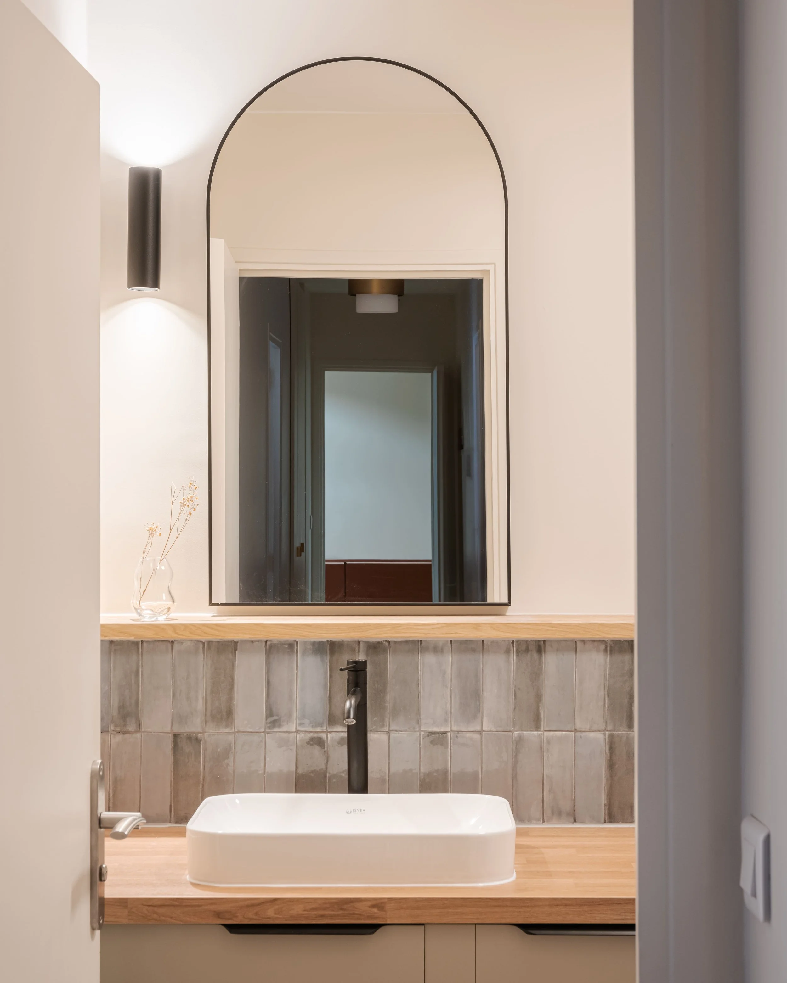 Salle de bain moderne avec un miroir, un lavabo blanc, un robinet noir, un éclairage mural et des carreaux gris derrière le lavabo.