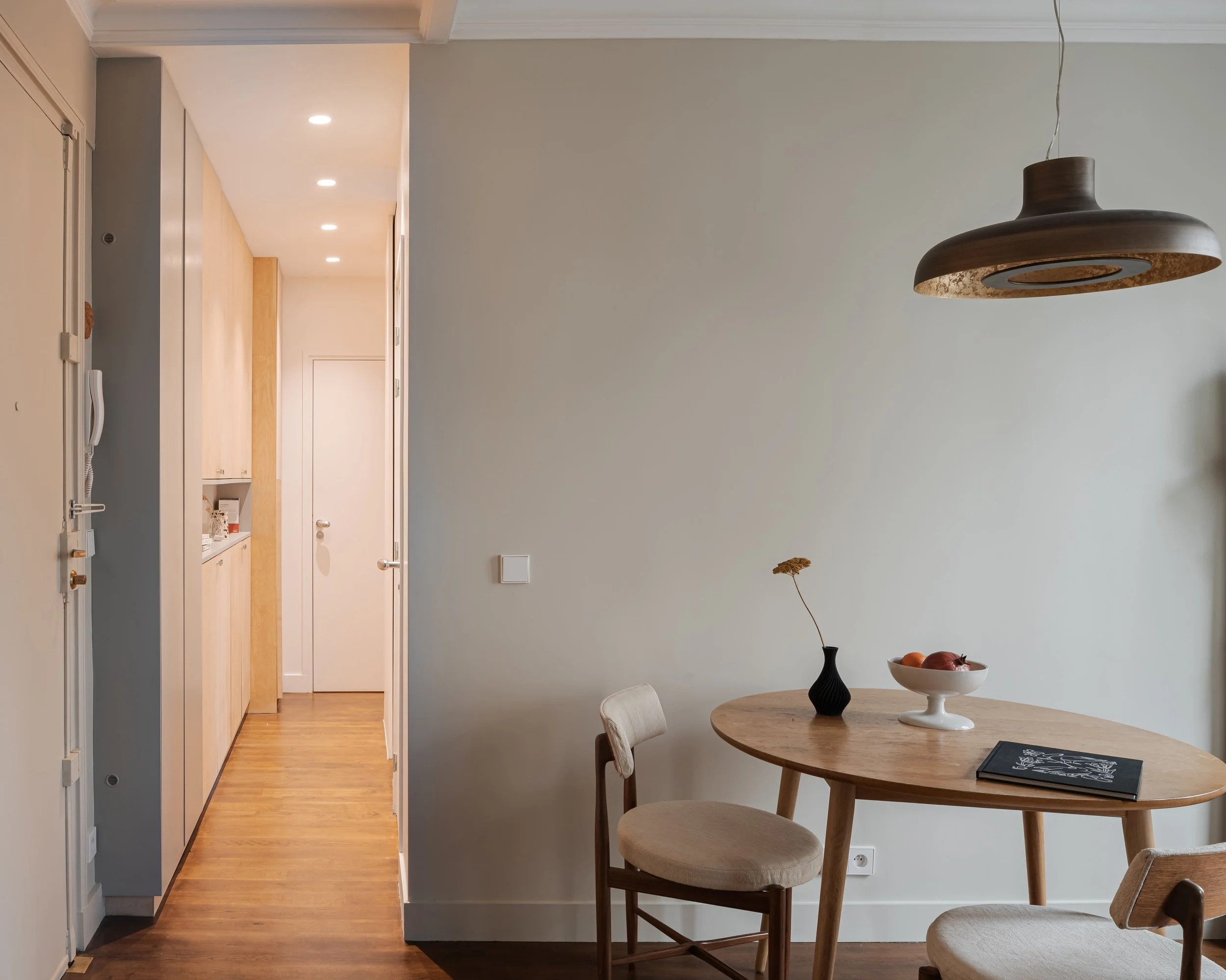 Intérieur d'une pièce avec une table en bois, deux chaises, un vase avec une fleur, un bol de fruits, et un plafonnier en bois. Un couloir menant à une porte blanche de l'autre côté.
