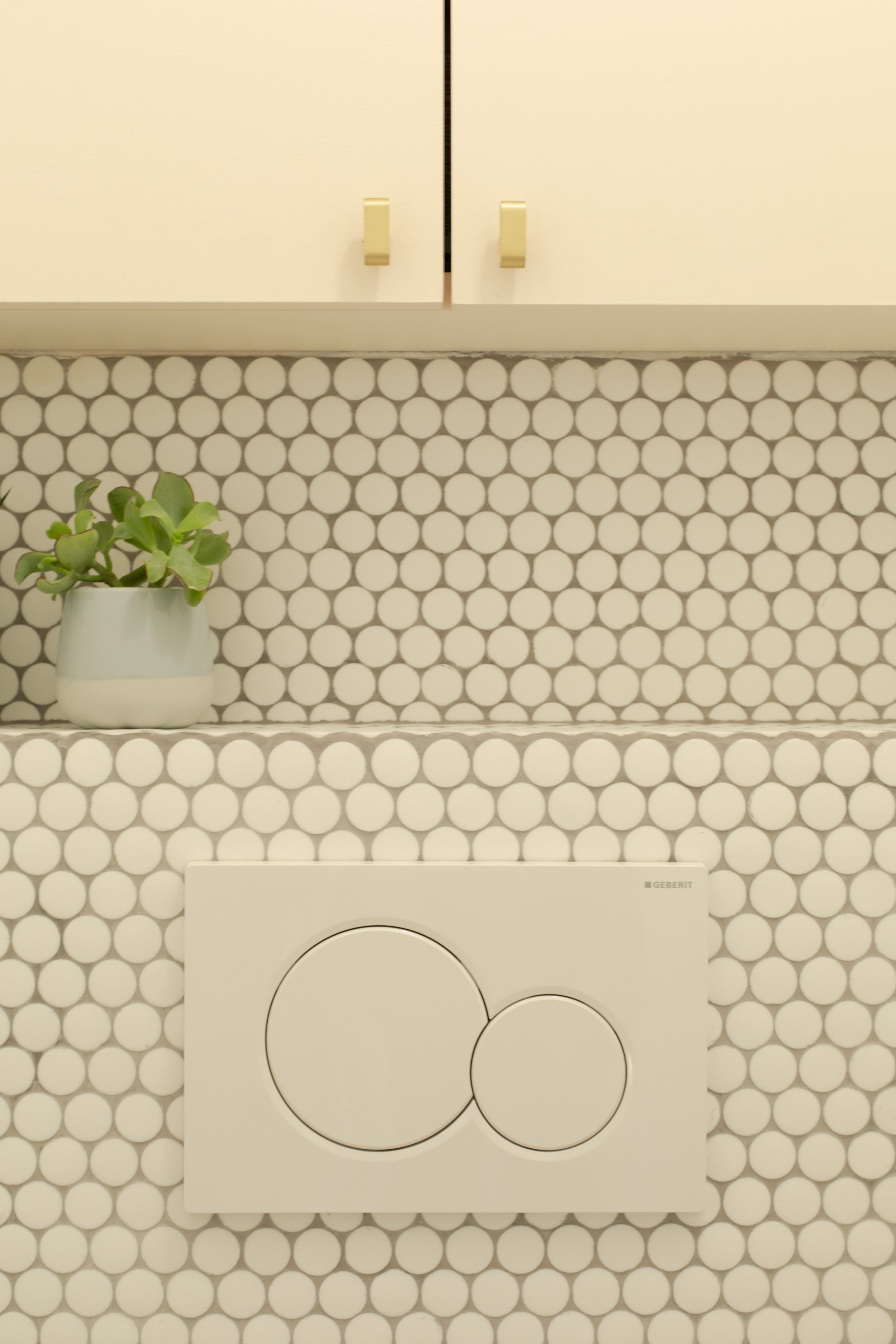 Une toilette suspendue encastrée dans un mur recouvert de carreaux de céramique blanc à motifs de cercles gris, avec une petite plante verte dans un vase blanc sur une étagère au-dessus.