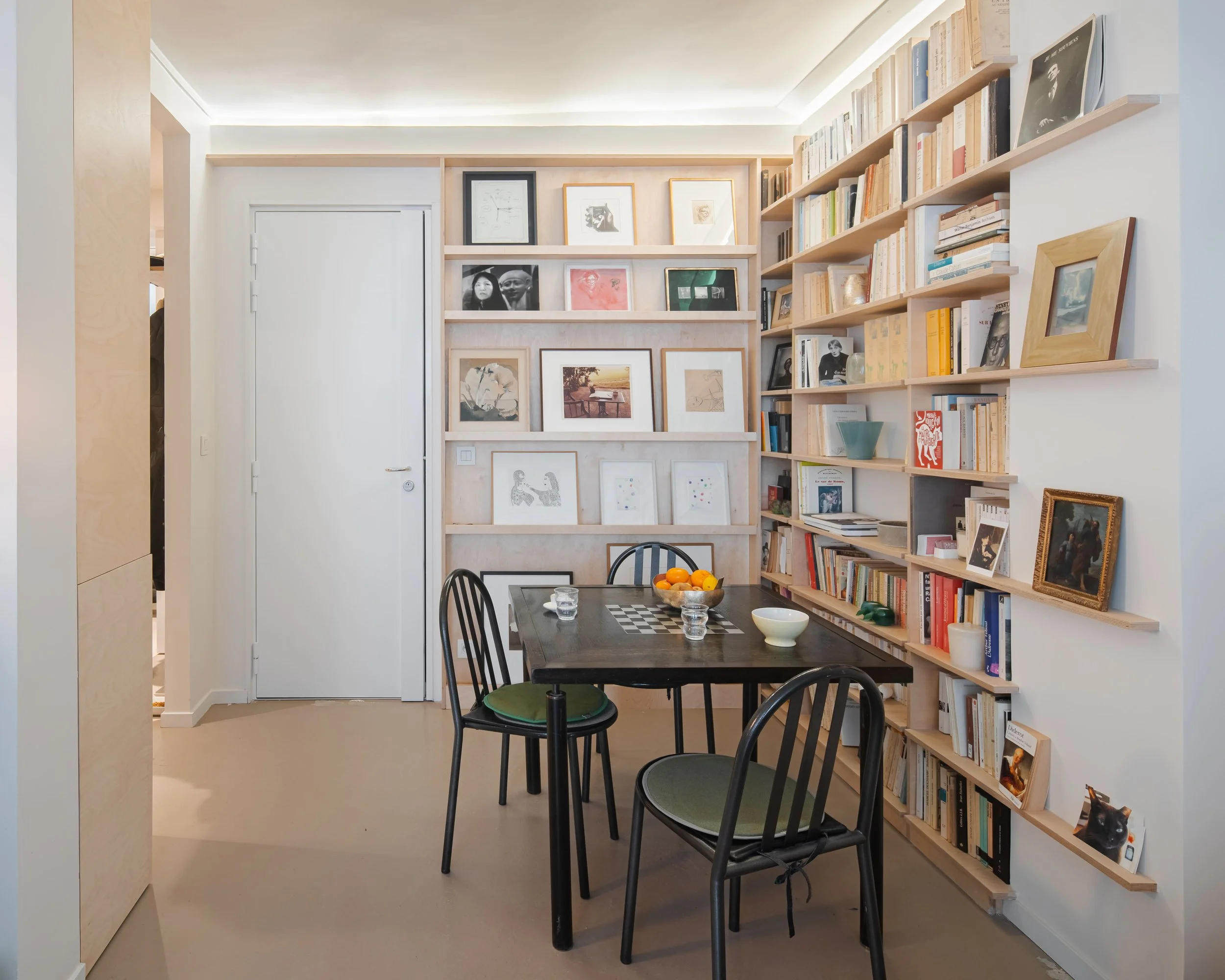 Salle à manger avec une table en bois foncé entourée de trois chaises noires avec des coussins verts. Sur la table, un bol de fruits, un bol blanc, deux verres et un porte-échecs. Murs blancs ornés d'étagères en bois clair remplies de livres et de ca