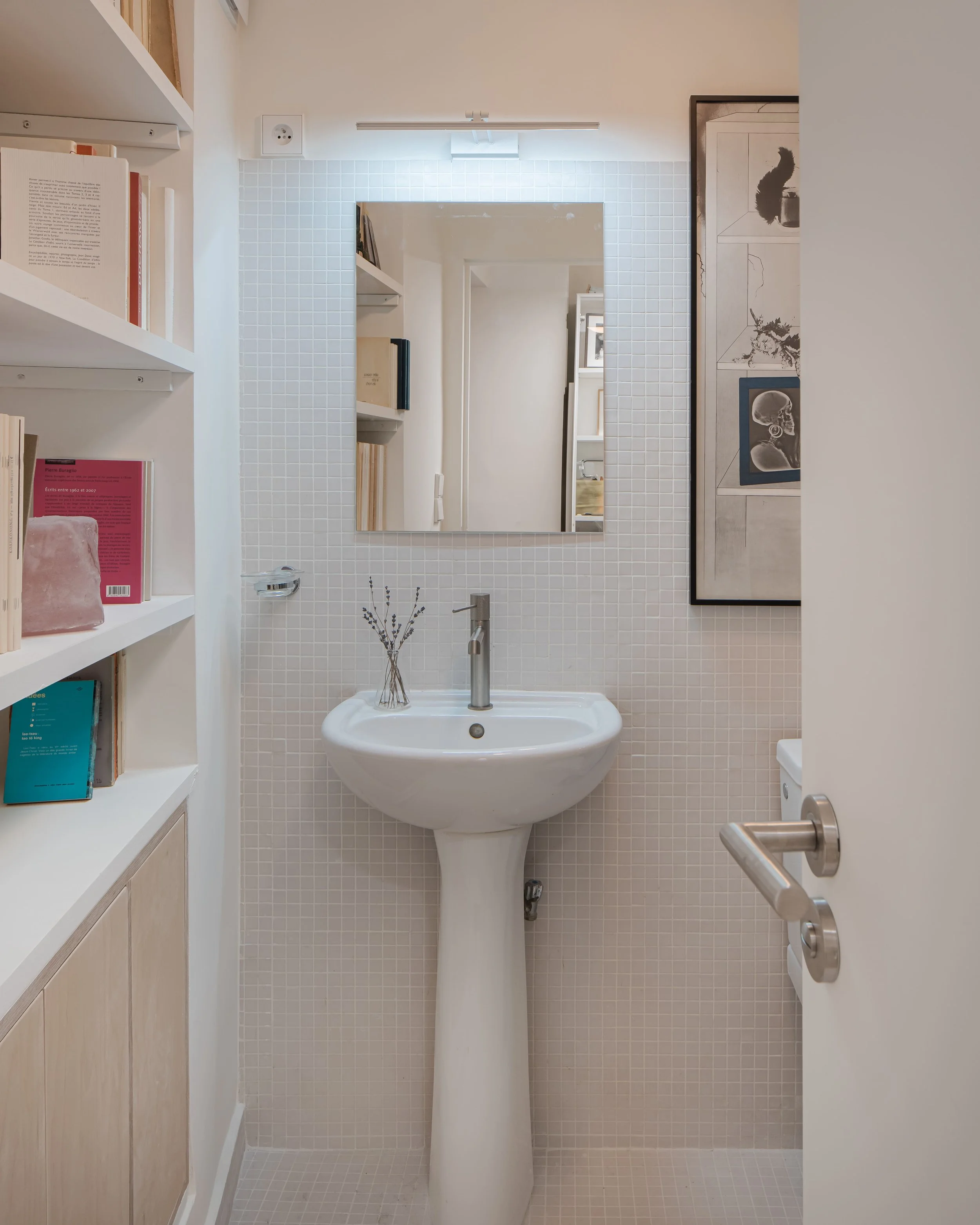 Petite salle de bain avec un lavabo blanc, un miroir carré, une étagère avec des livres et une décoration en lavande dans un vase. Mur en carrelage blanc et décoration murale artistique à droite.