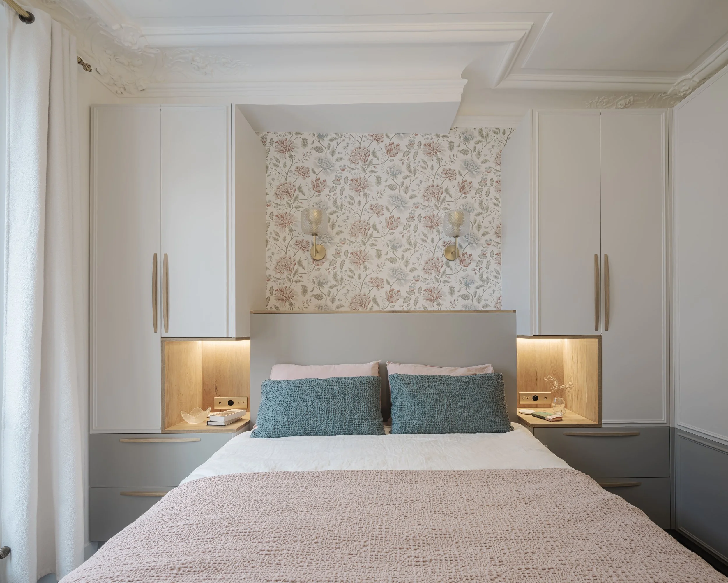 Chambre avec lit double, deux oreillers bleu et couette beige, panneaux de rangement gris avec éclairage, toile de fond de papier peint floral, appliques murales, rideaux blancs.