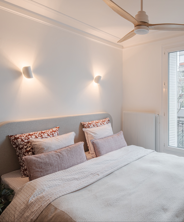 Chambre à coucher avec un lit, oreillers, mur lumineux, fenêtre et ventilateur au plafond.