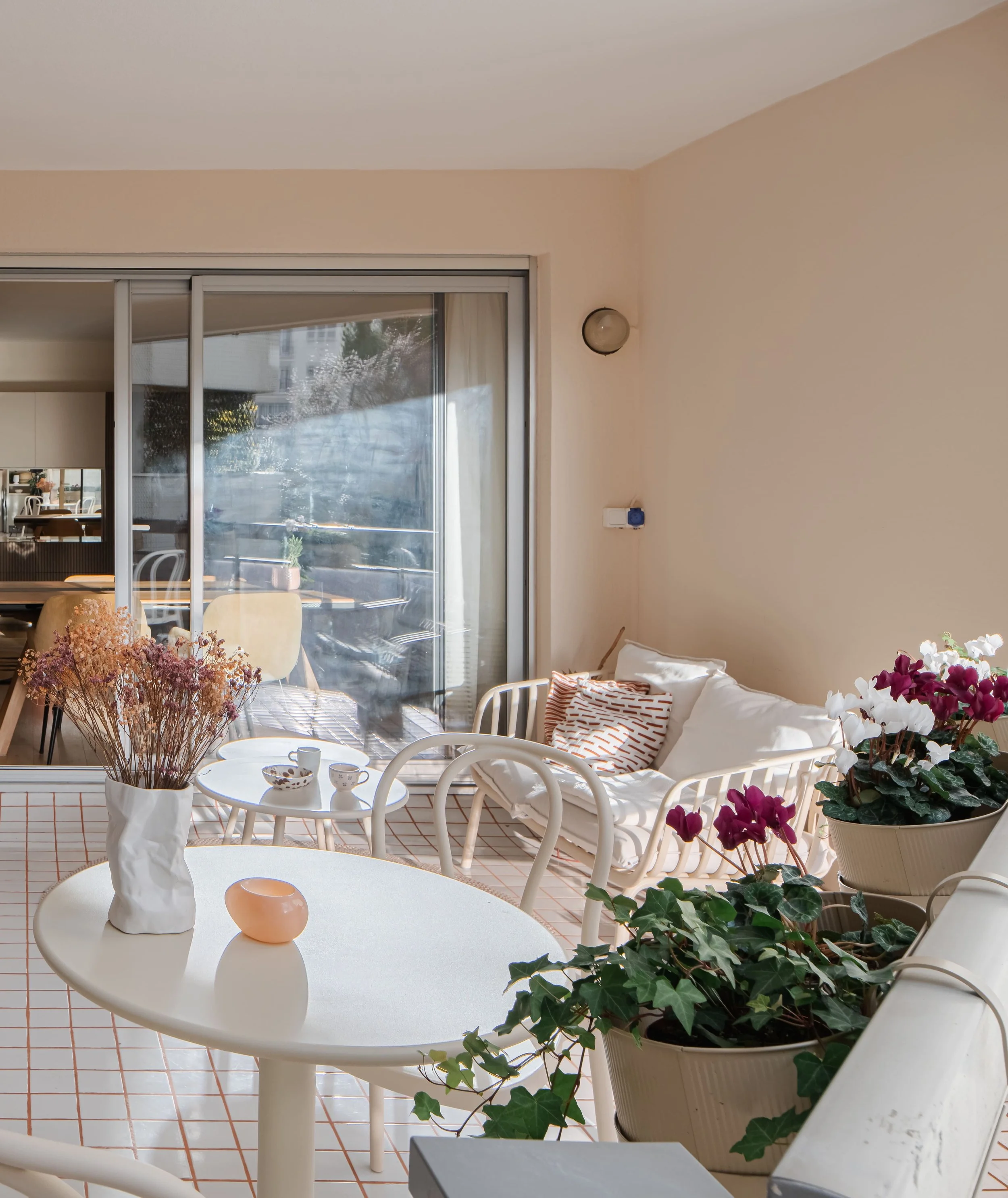 Balcon ensoleillé avec une table ronde, des vases de fleurs, des chaises blanches, un canapé avec des coussins, et des plantes en pot, avec une porte-fenêtre donnant sur un espace extérieur.