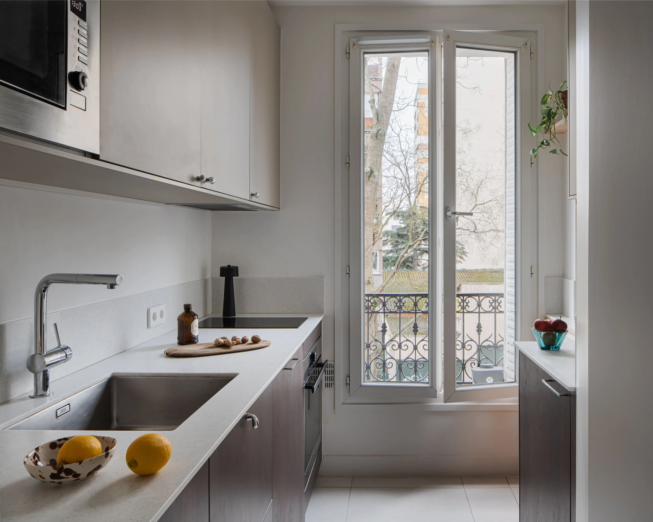 Cuisine moderne avec fenêtre donnant sur un balcon en fer forgé, meuble de cuisine en bois clair, évier à gauche, quelques citrons dans un bol, fruits rouges dans un bol en verre bleu à droite, plantes suspendues