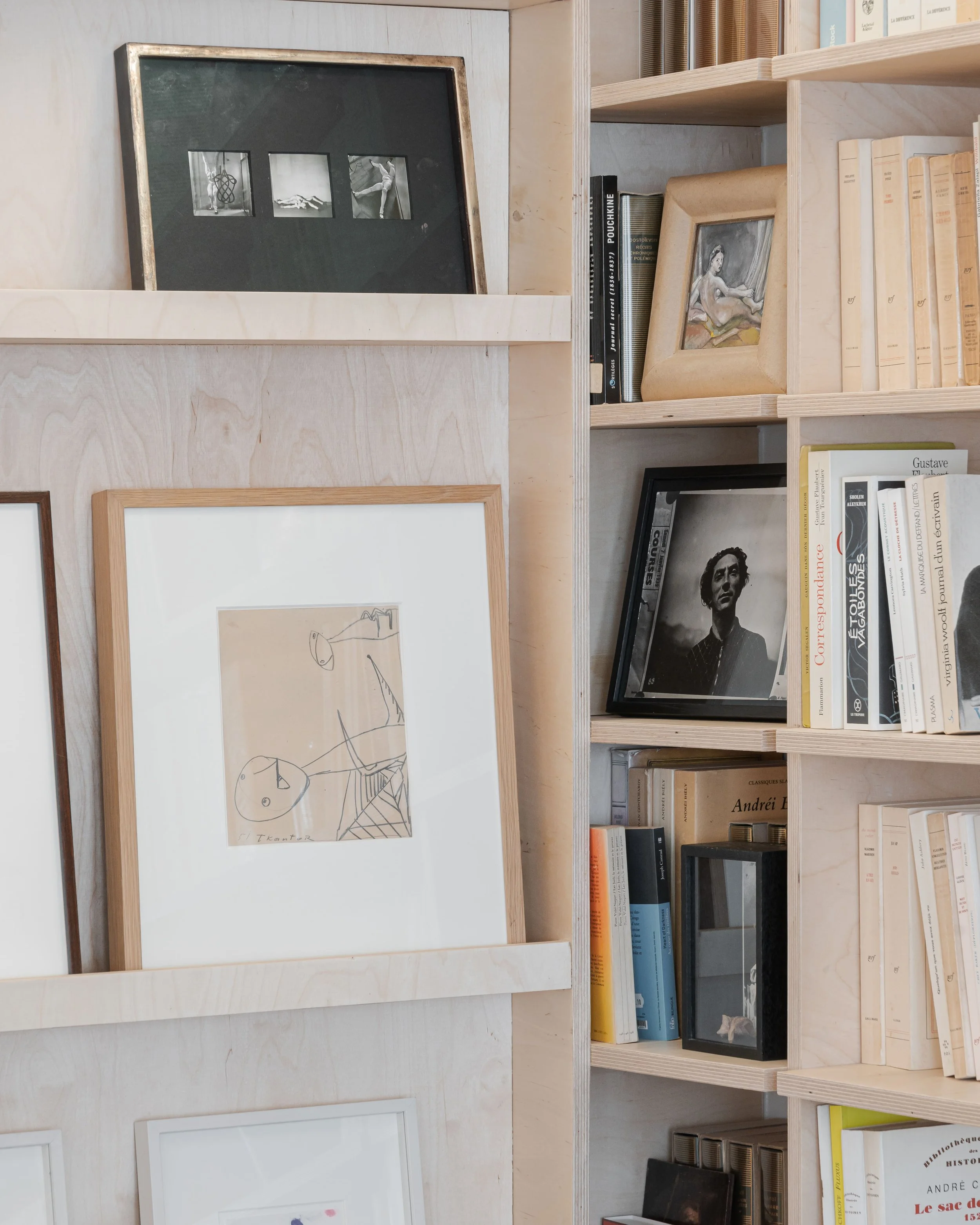 Une étagère en bois avec plusieurs livres, premières pages visibles, accompagnée de photos encadrées, notamment une photo noir et blanc d'une femme et un tableau d'un dessin minimaliste d'une personne.