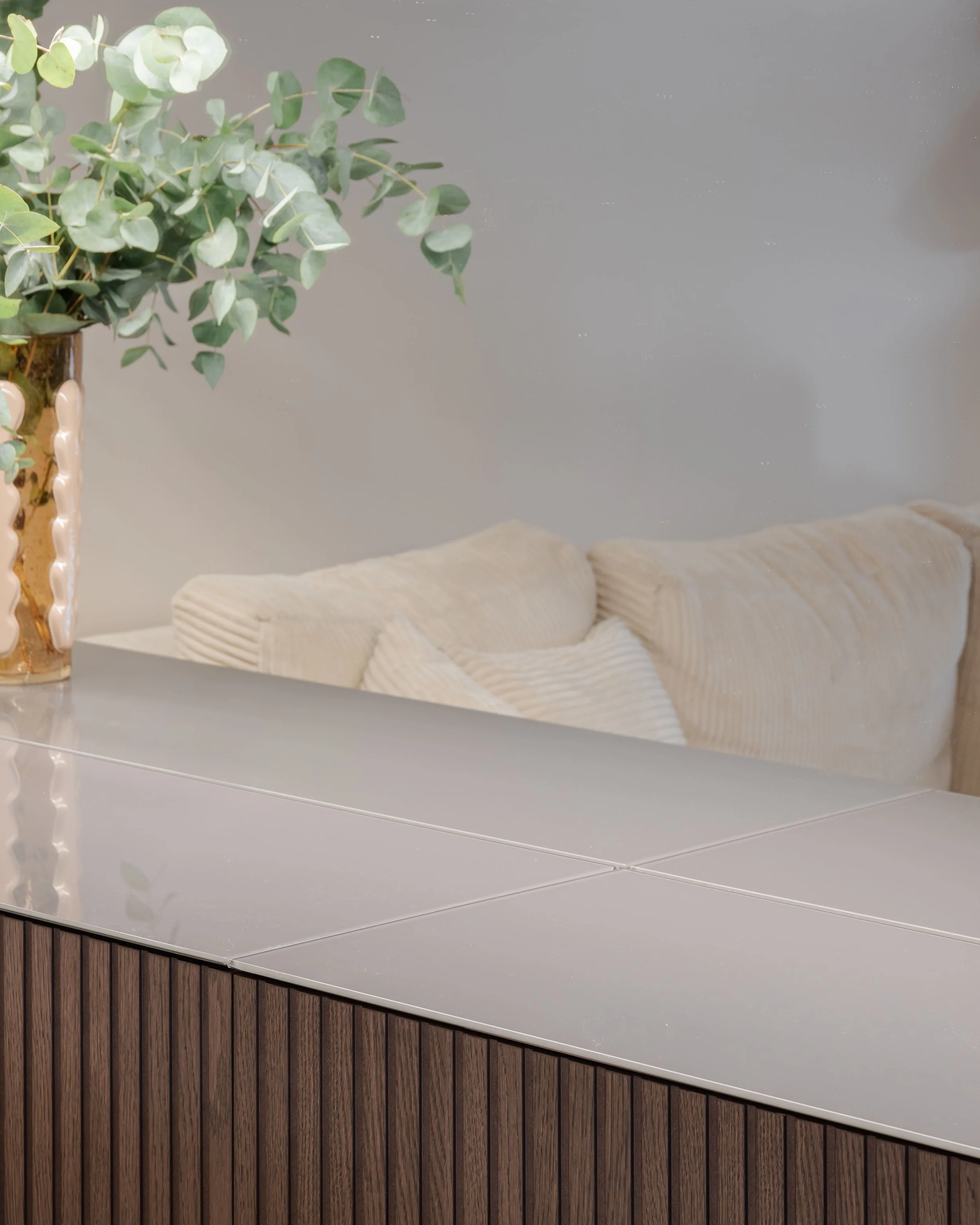 Vase avec des feuilles de eucalyptus sur une surface en bois et un canapé beige avec des coussins en arrière-plan.