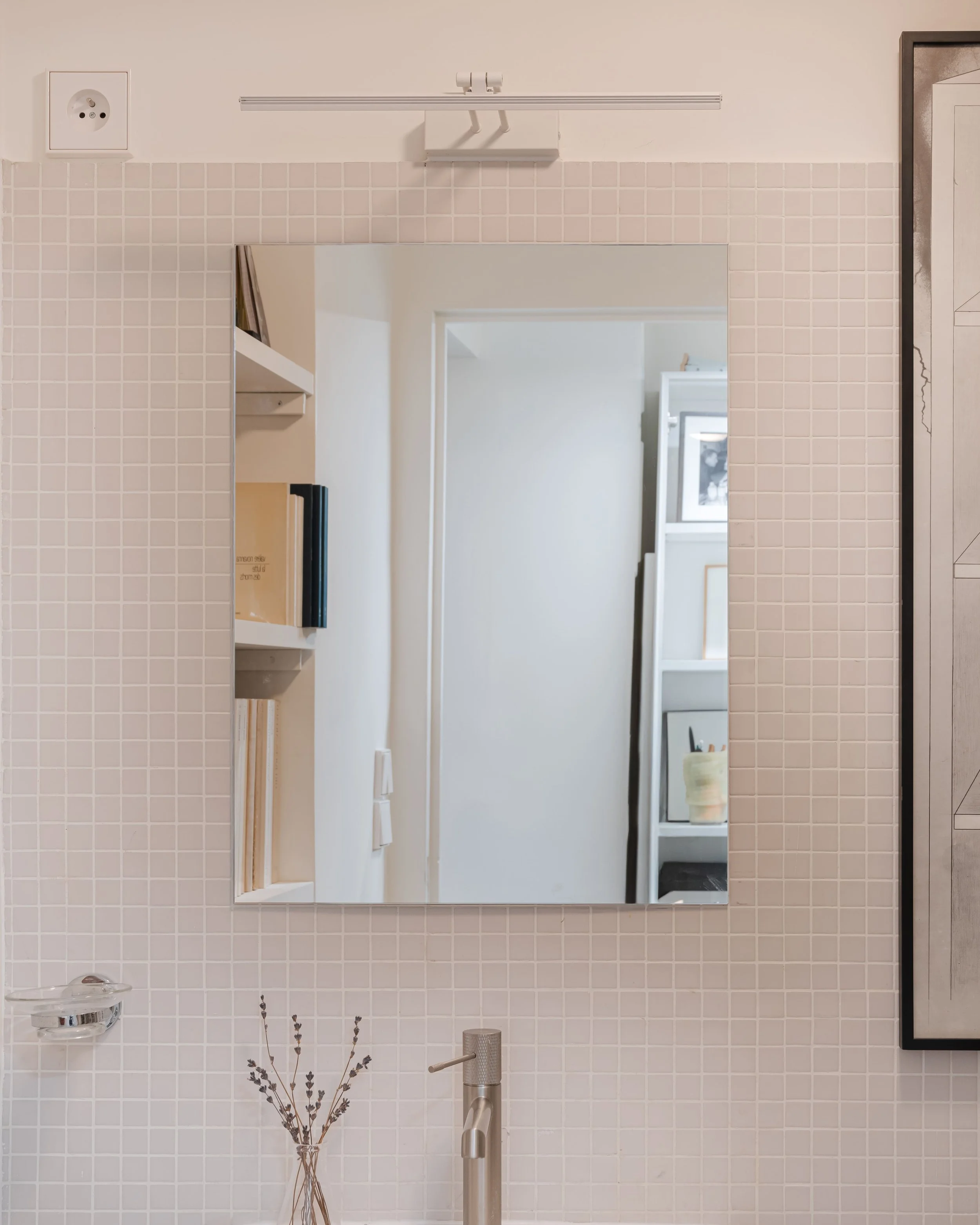 Miroir au-dessus d'un évier dans une salle de bain avec carrelage beige, une étagère en haut, une prise électrique, et des étagères avec des livres et une boîte