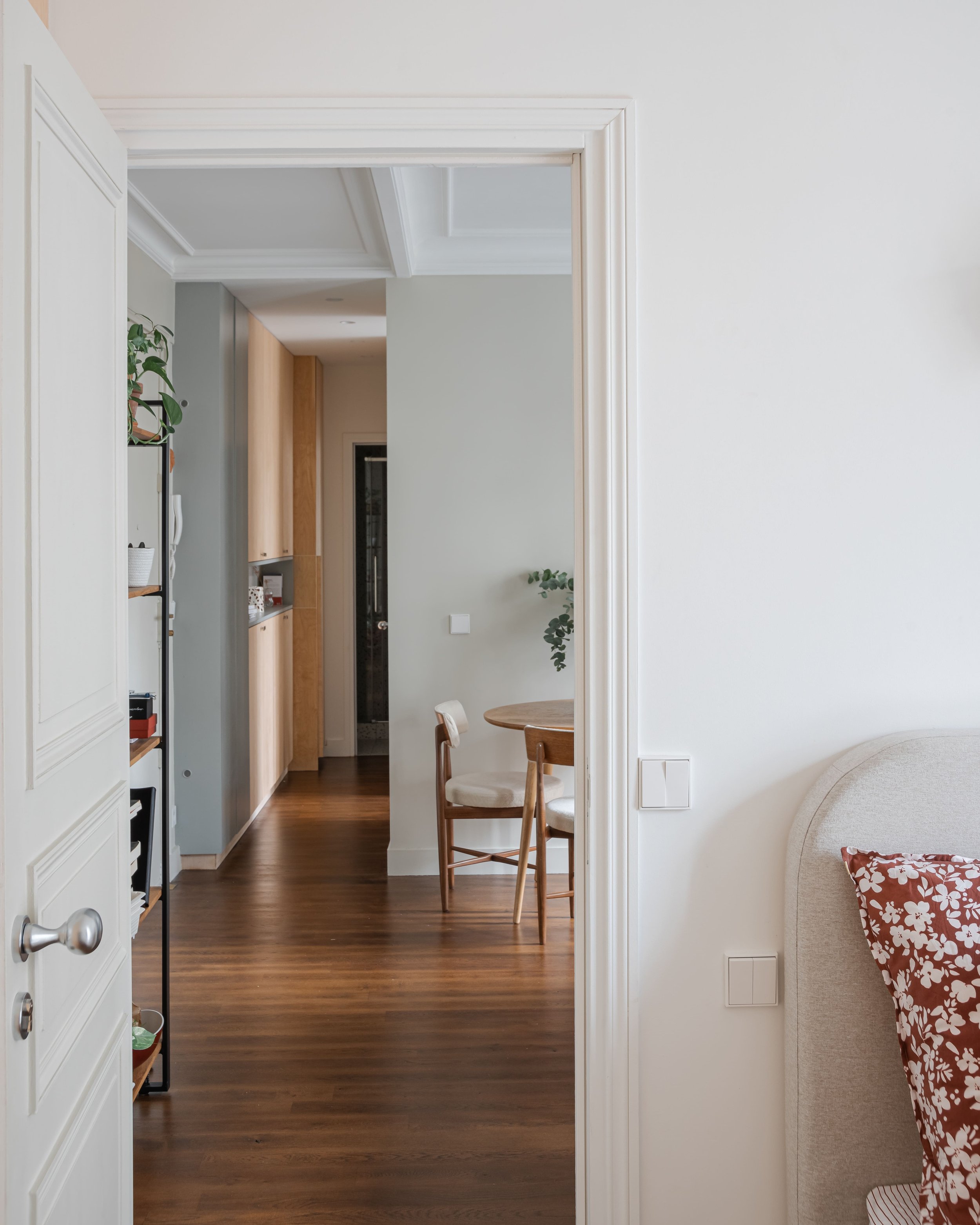 Intérieur d'une maison avec porte ouverte sur un espace de vie comprenant un mobilier en bois, une table ronde, des chaises, un lit avec un coussin floral, et un mur blanc.