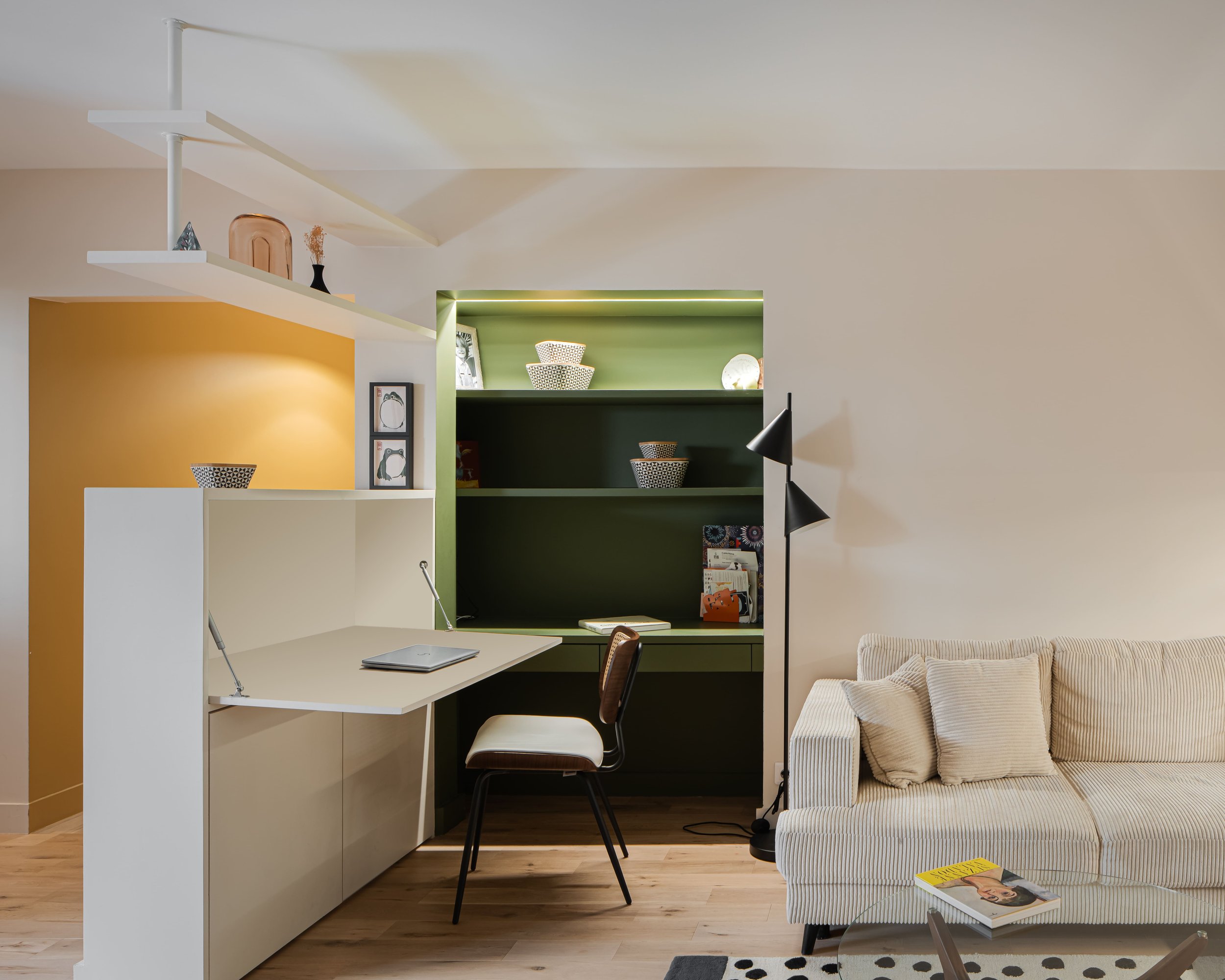 Intérieur d'un salon avec un espace de bureau, étagère verte avec des livres et des vases, canapé beige rayé avec des coussins, lampe sur pied et table avec magazine.