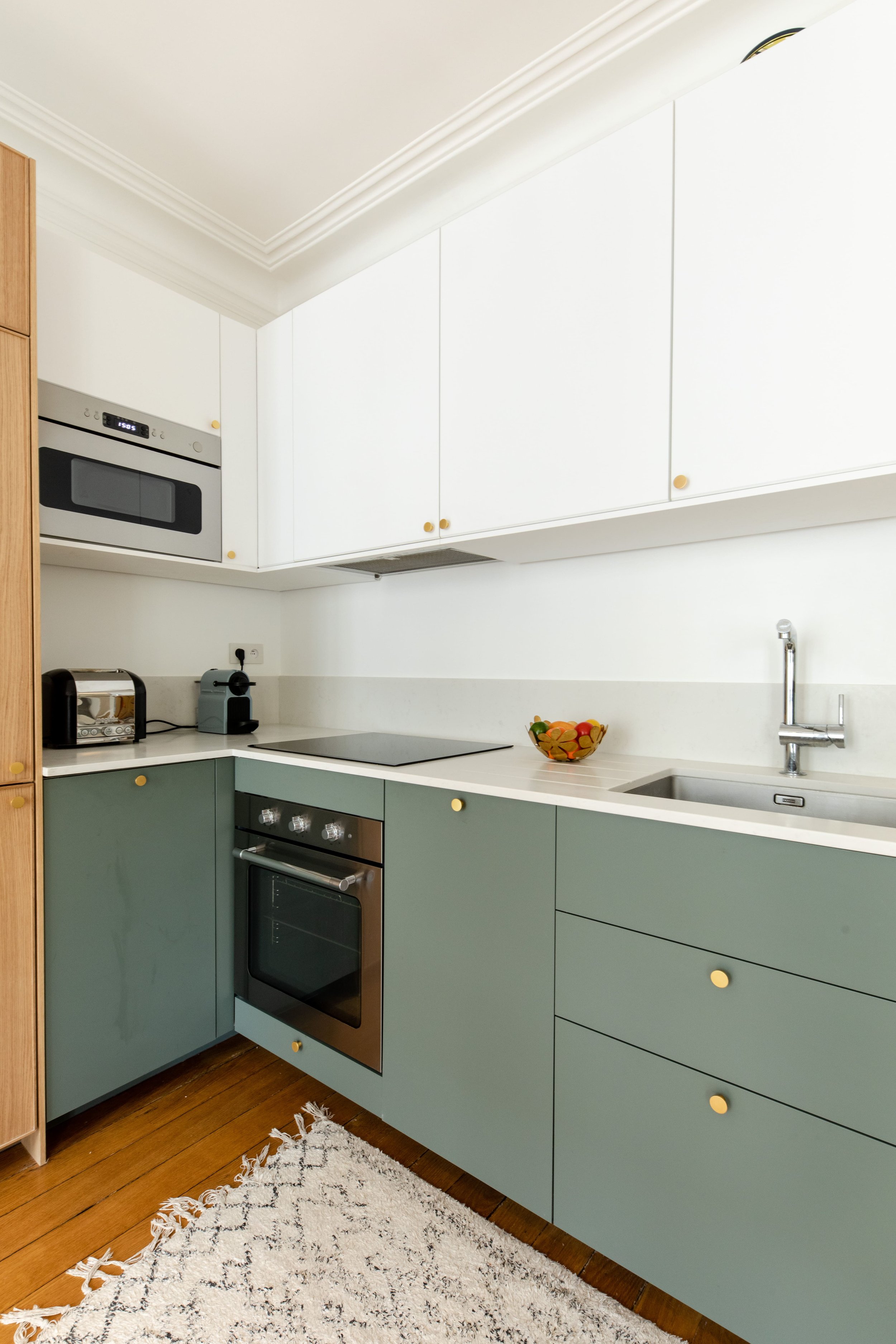 Cuisine moderne avec four, plaque de cuisson, micro-ondes, grille-pain, machine à café, évier, bol de fruits, et placards blancs et verts avec poignées jaunes.