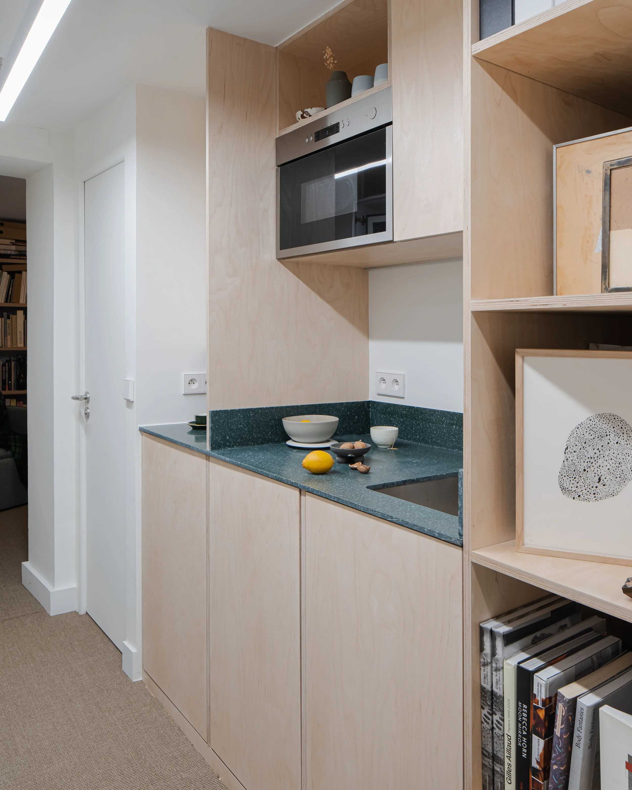 Cuisine moderne avec plan countertop en pierre verte, containing des bowls et fruits, et meuble en bois clair avec étagères.
