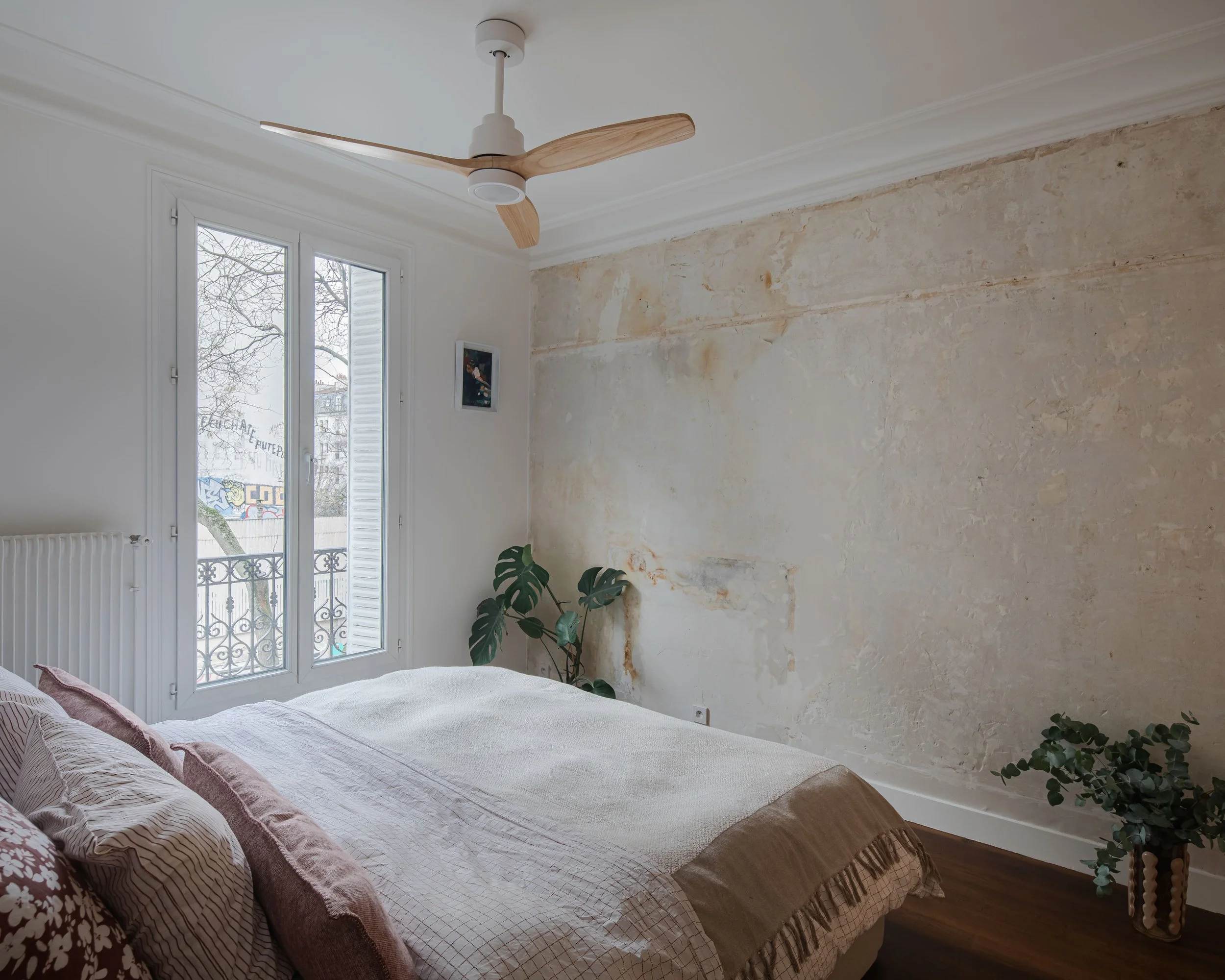 Chambre avec un lit blanc, plante verte, fenêtre avec vue sur la ville, mur en cours de rénovation, ventilateur au plafond.