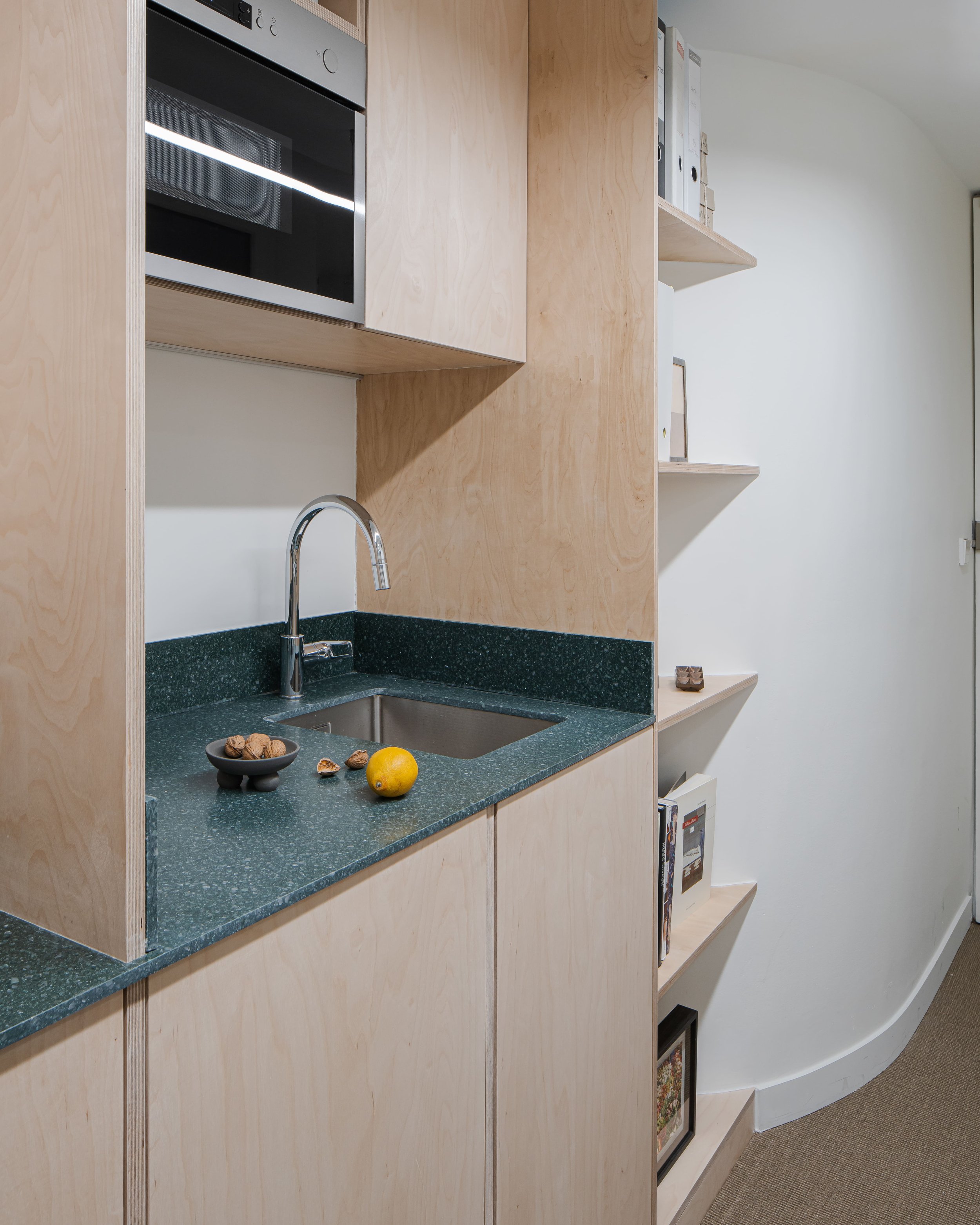 Coin cuisine avec évier, robinet, micro-ondes, étagères en bois avec livres et cadres, et fruits sur le plan de travail.