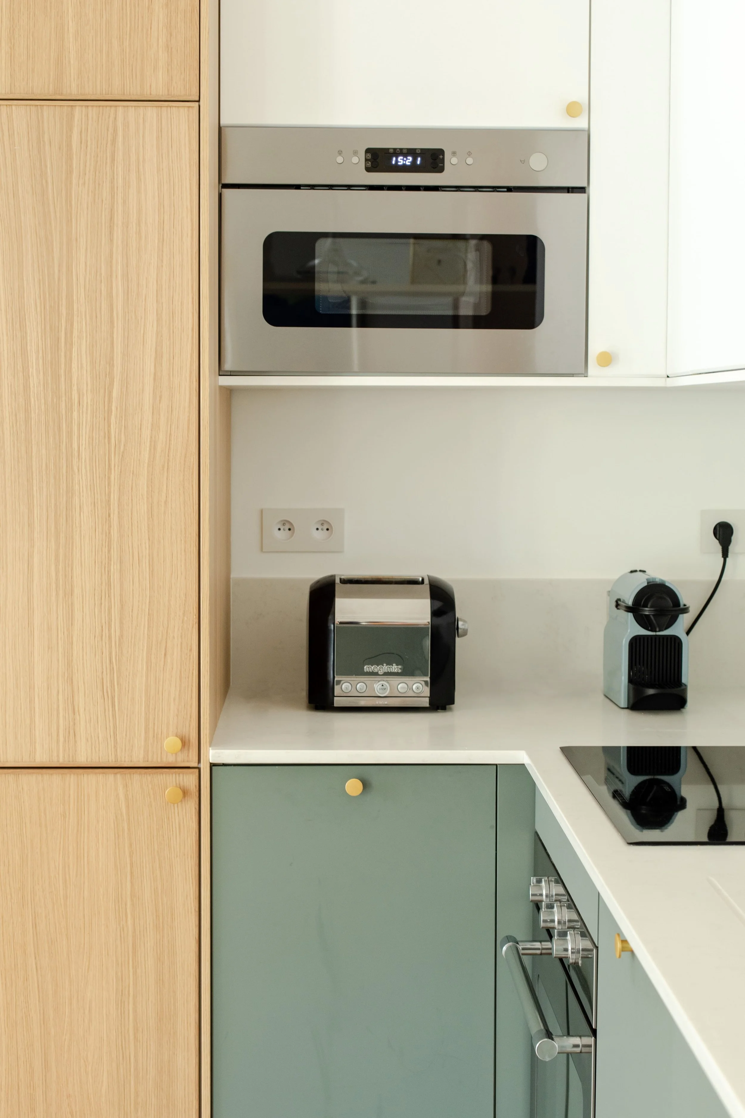 Cuisine moderne avec un four encastré, une cafetière à capsules, un grille-pain noir, et une plaque à induction sur un plan de travail blanc, avec des armoires vertes et en bois.