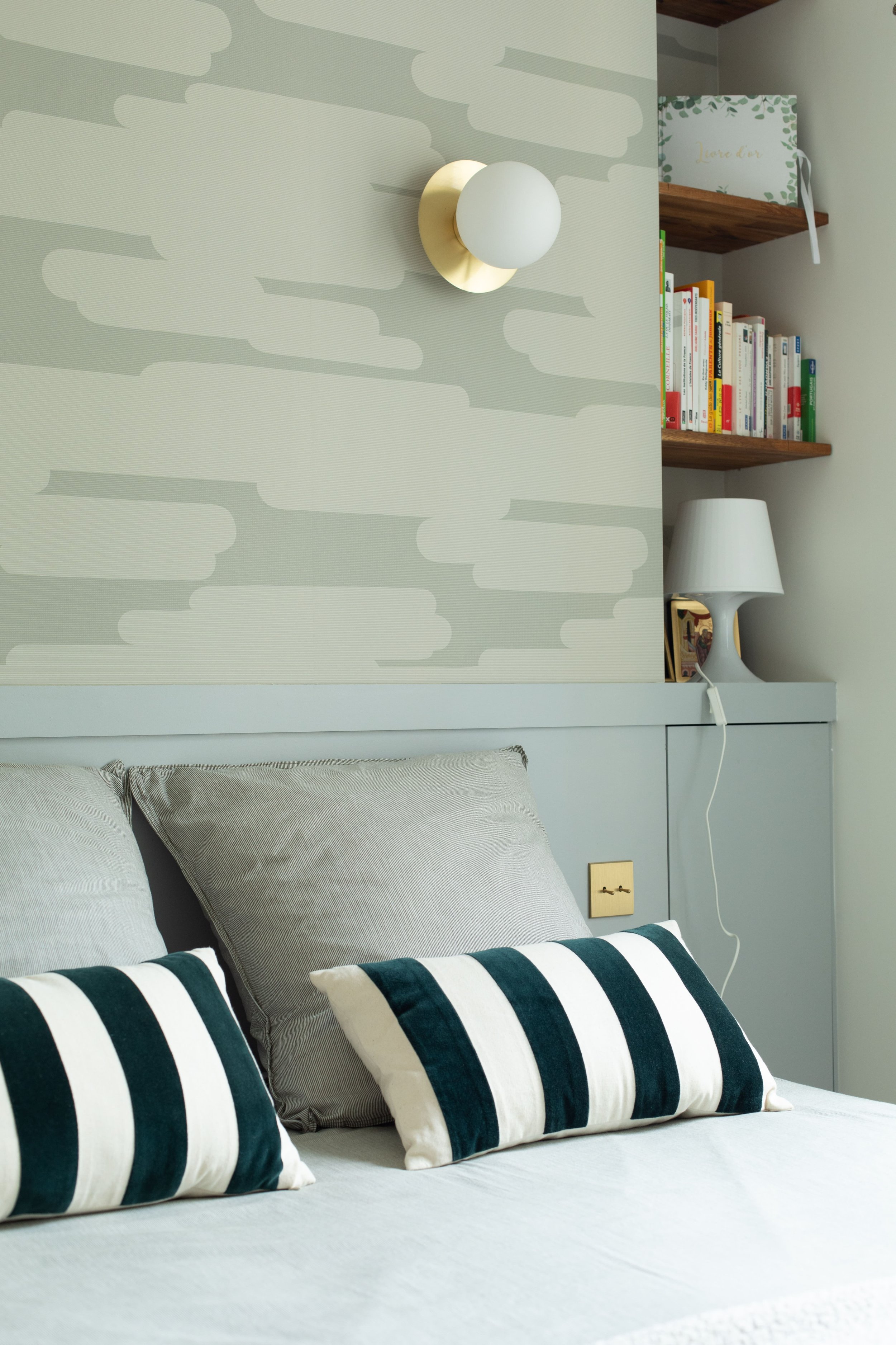 Chambre avec lit,oreillers gris et à rayures, lampe de chevet blanche, étagères en bois avec des livres et une boîte décorative.