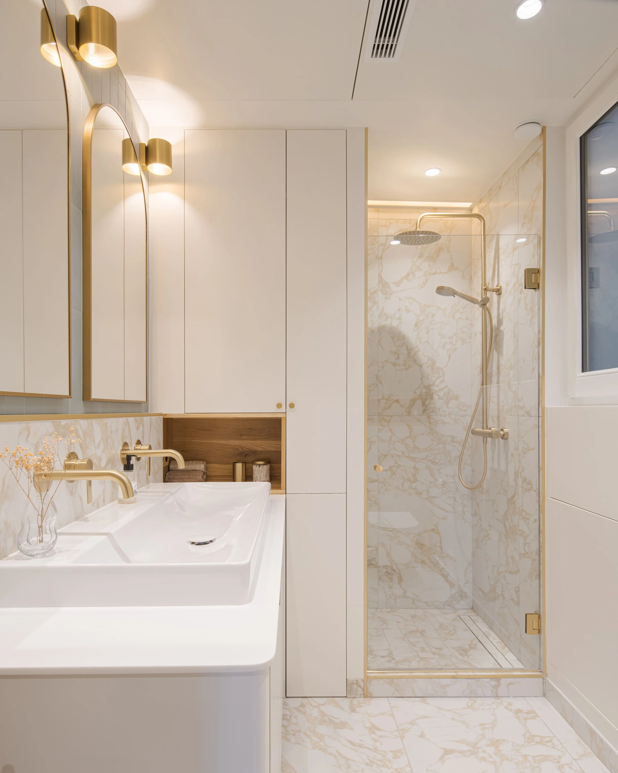 Salle de bain élégante avec vasque blanche, miroirs avec encadrement doré, éclairage mural doré, placards blancs, cabine de douche en marbre beige avec détails dorés, et une fenêtre offrant de la lumière naturelle.