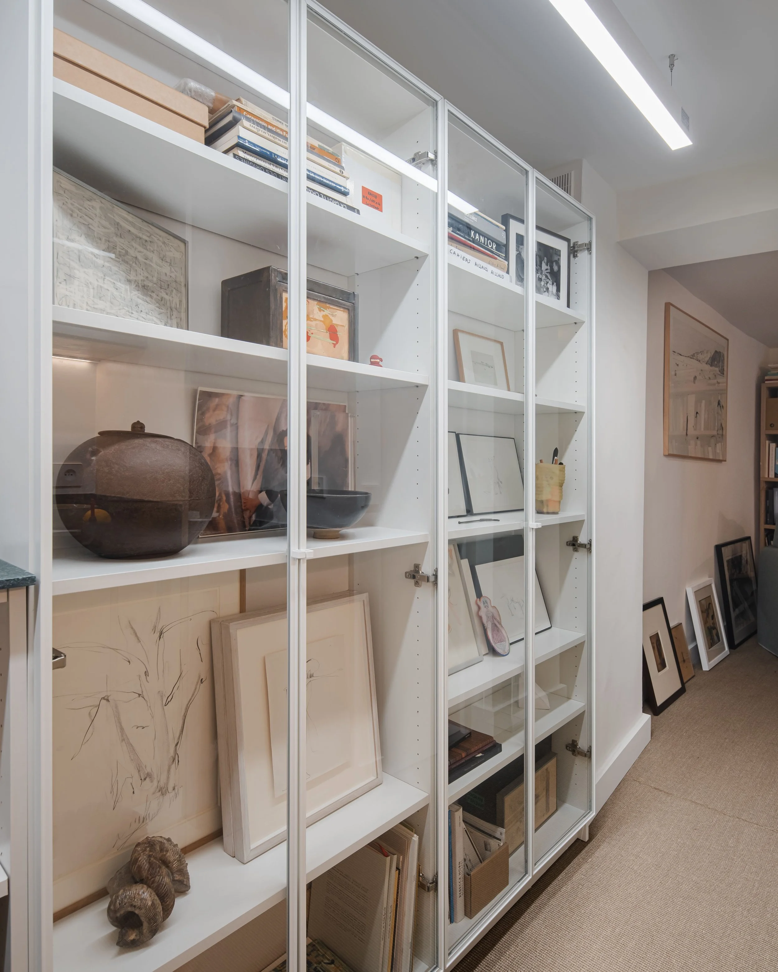 Étagère blanche contenant des livres, des cadres, des objets décoratifs et des dessins dans une pièce bien éclairée.