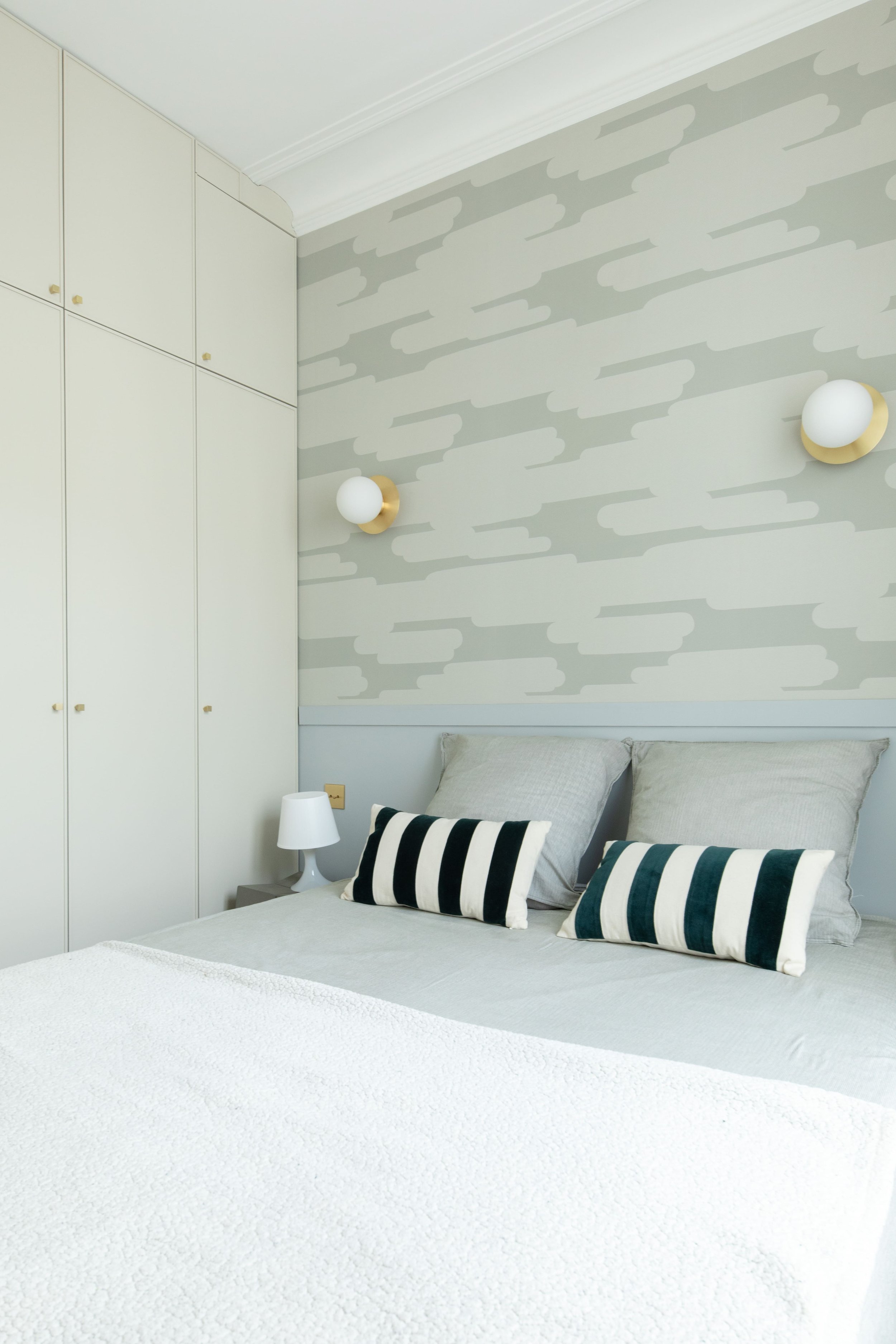 Chambre à coucher avec un lit, deux oreillers rayés noirs et blancs, deux autres oreillers gris, une table de chevet avec une lampe blanche, un mur avec un motif abstrait gris et blanc, deux appliques murales avec globes blancs.