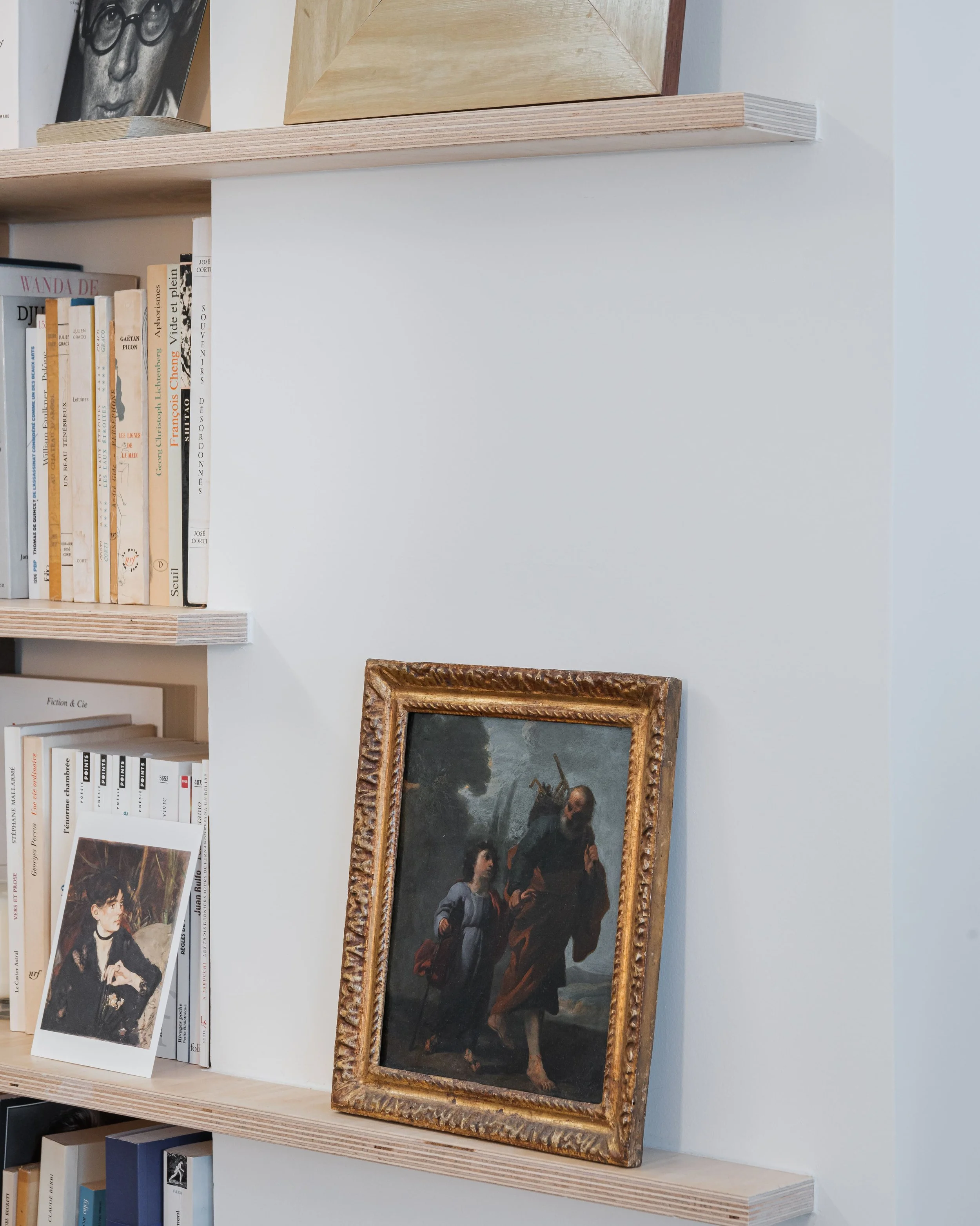 Une étagère en bois avec des livres alignés verticalement et une photographie en noir et blanc d'un homme portant des lunettes, ainsi qu'une peinture encadrée représentant deux personnages, dont l'un porte une arme sur l'épaule.