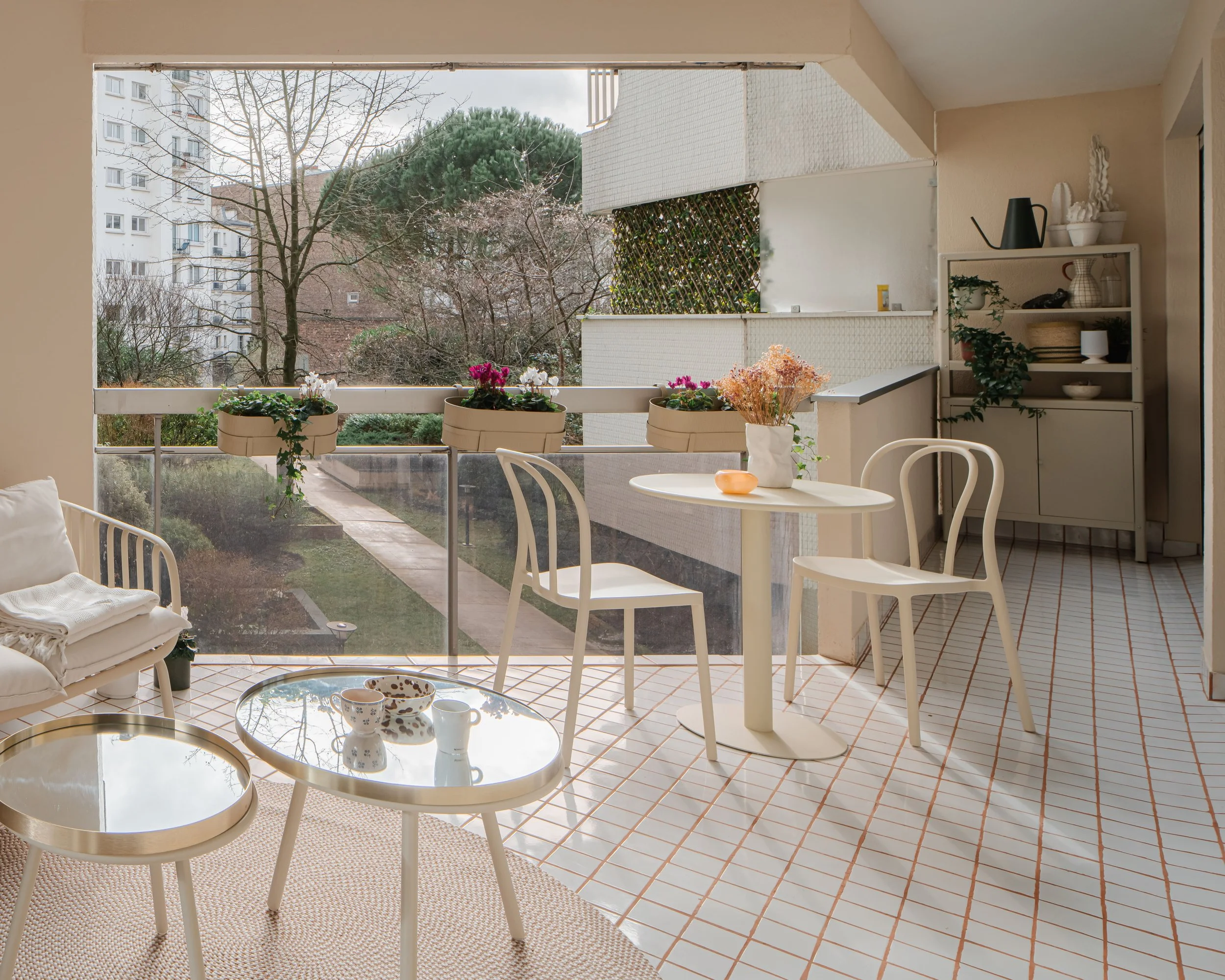 Balcon avec mobilier blanc, fleurs en pot, vue sur la ville et arbres, carrelage blanc et terrasses modernes.